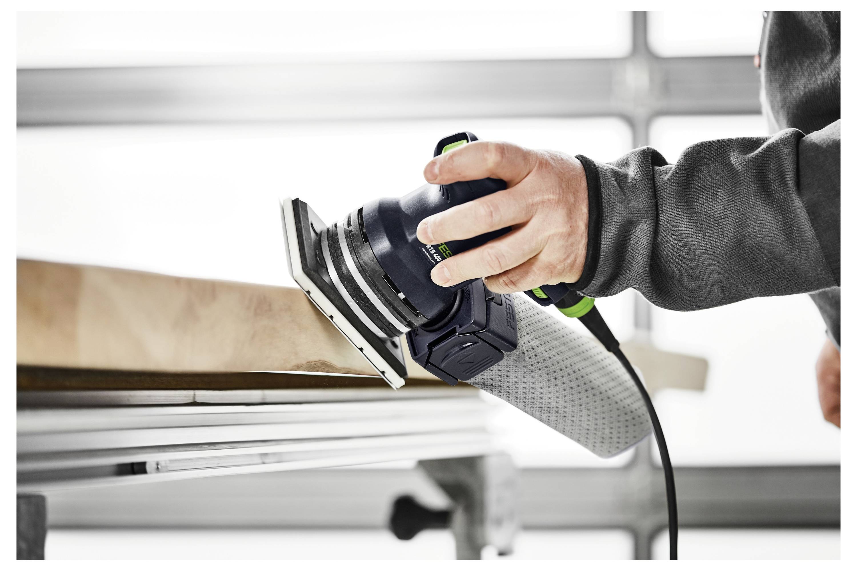 Une personne utilise une ponceuse électrique sur une surface en bois, illustrant le travail du bois ou la fabrication de meubles dans un environnement d'atelier.