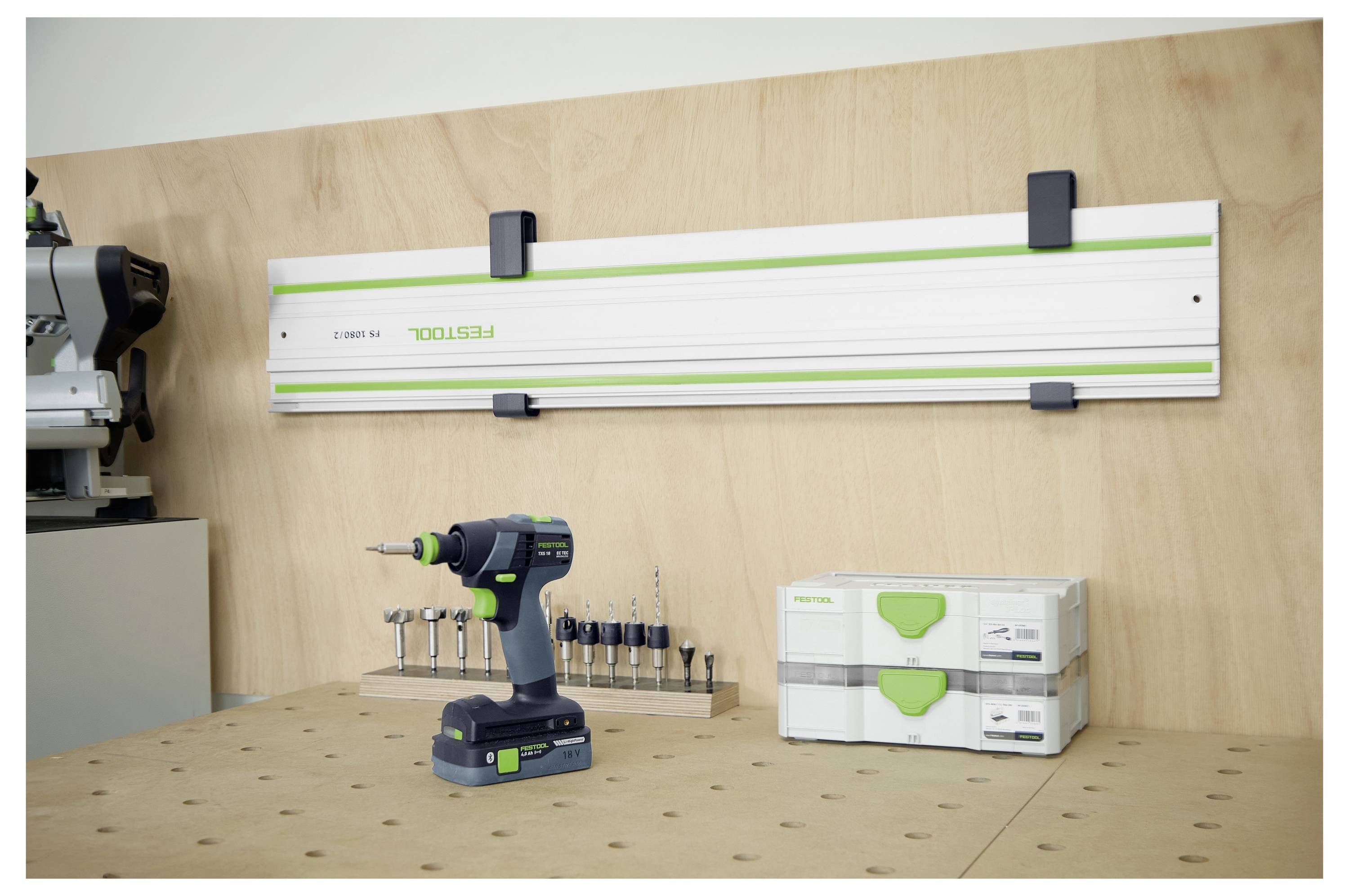 Un espace d'atelier présentant une perceuse électrique avec des mèches, un rail de guidage suspendu au mur, et des boîtes à outils empilées sur un établi en bois.