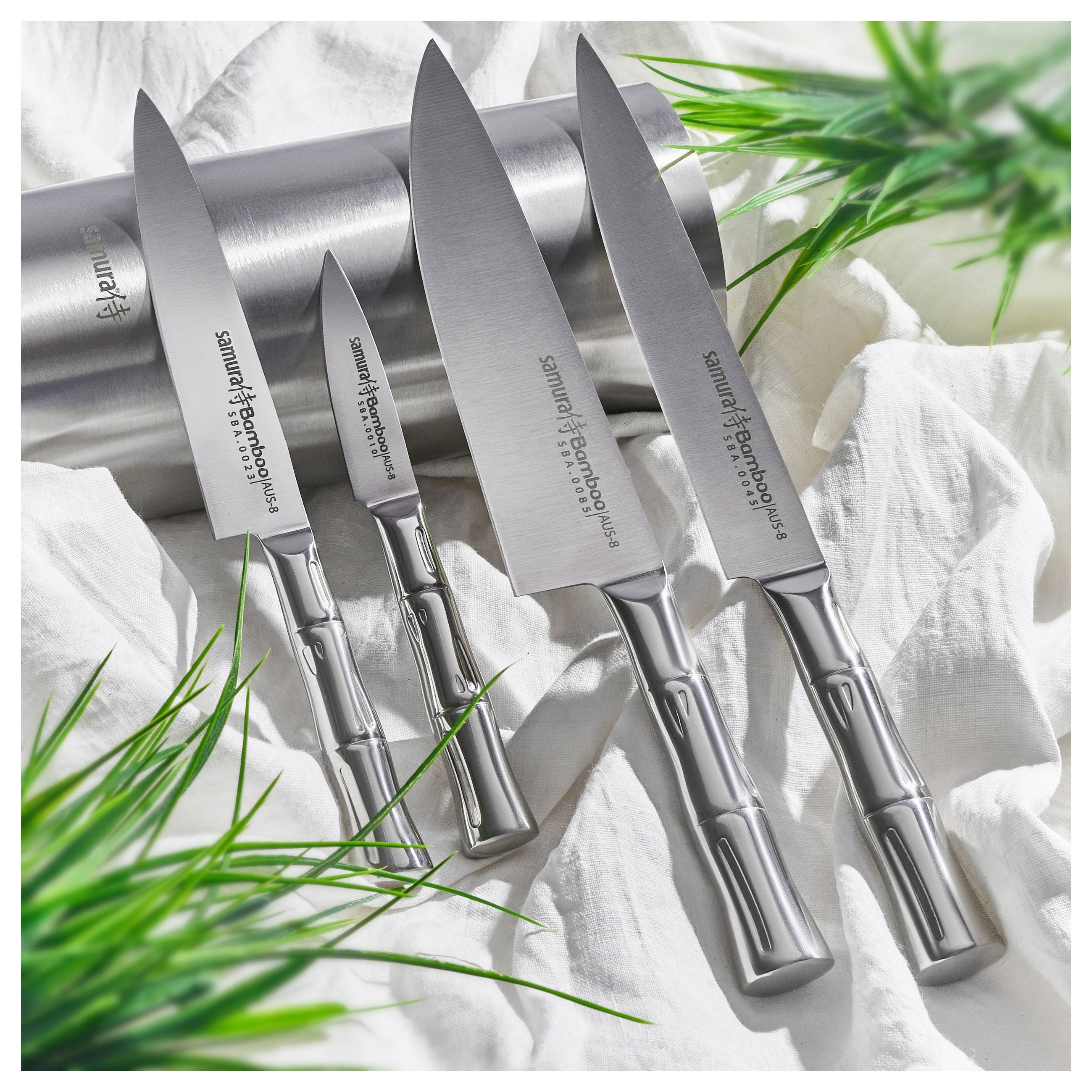 Four stainless steel knives with engraved handles are displayed alongside a cylindrical knife holder, surrounded by green leaves on a white cloth.