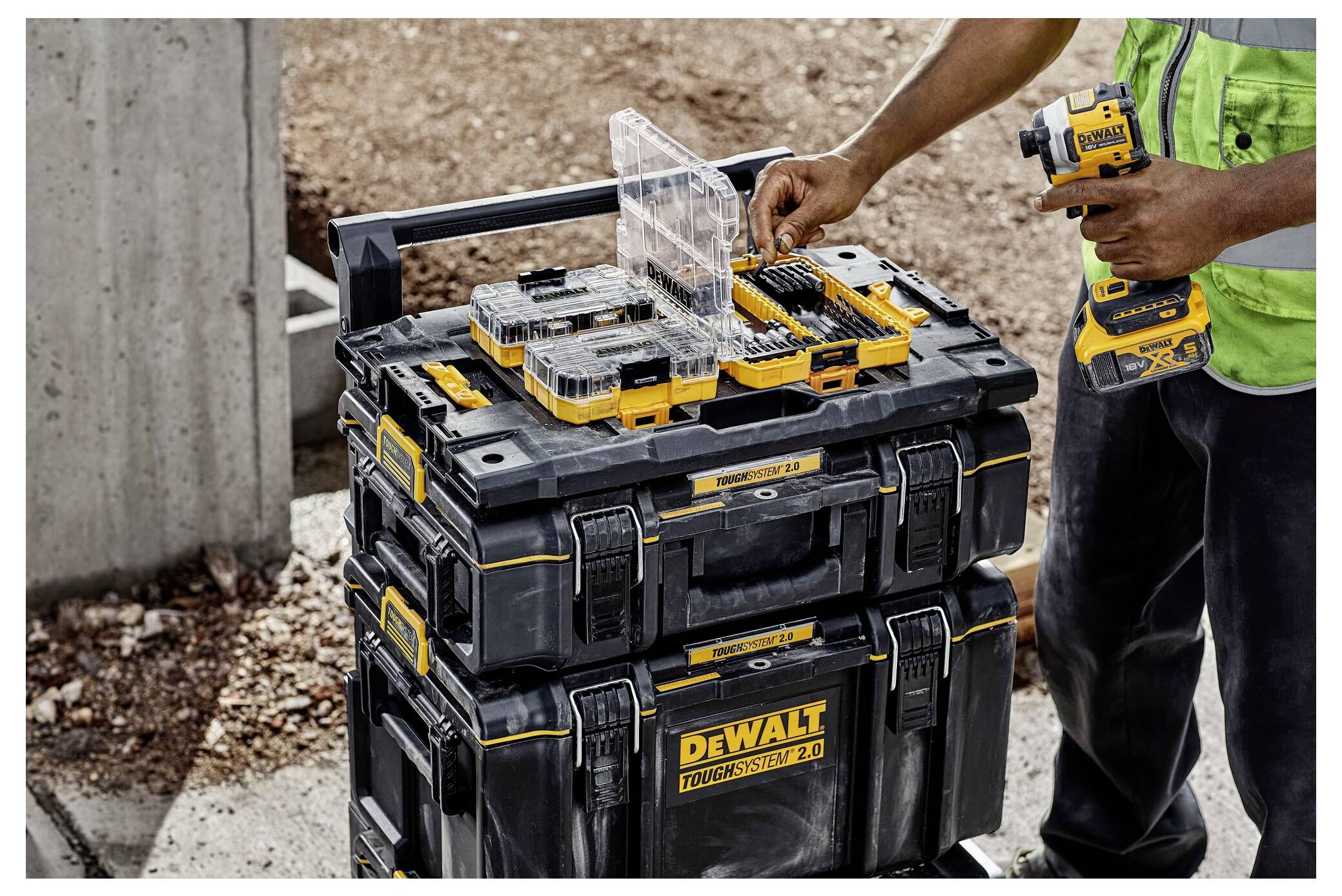 Un ouvrier organise des outils dans un coffre de rangement DEWALT noir et jaune, tenant une perceuse électrique. Le cadre semble être un chantier de construction.