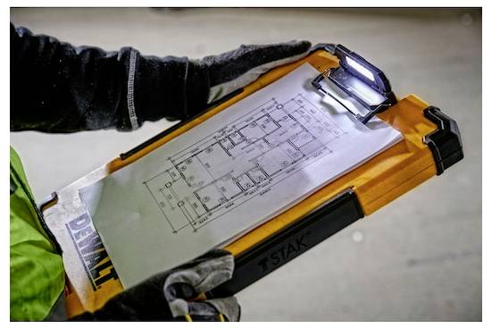 Une personne portant des gants tient un presse-papiers avec un document de plan d'étage attaché par une pince. Le presse-papiers a une lumière en haut.
