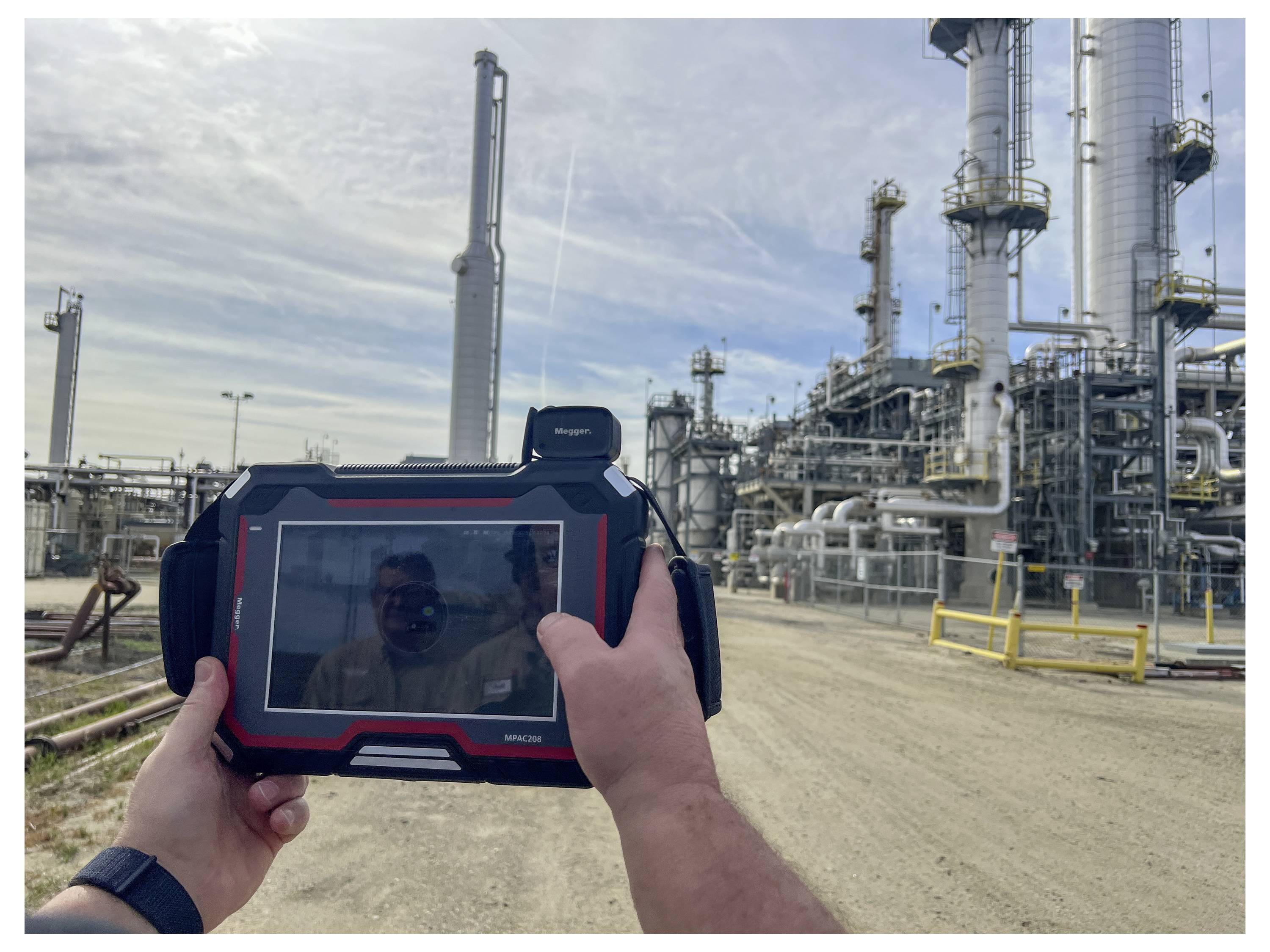 Une personne utilise une tablette pour contrôler ou inspecter une usine industrielle, avec des pipelines et des tours, probablement pour surveiller l'équipement ou la sécurité.
