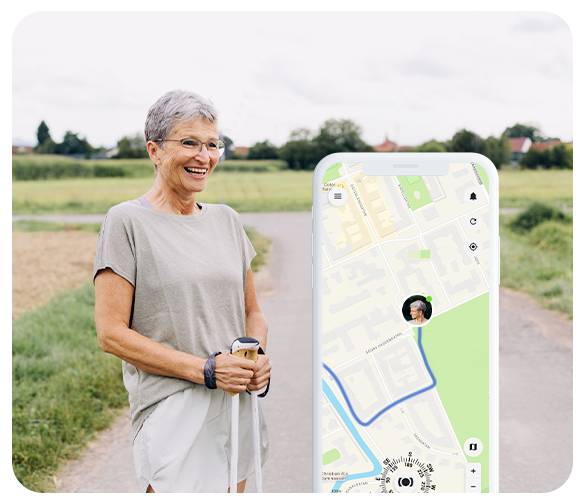 Une personne souriante aux cheveux gris courts, vêtue de vêtements décontractés, se tient debout sur un sentier de campagne avec des bâtons de marche. Un téléphone affiche une application de carte.