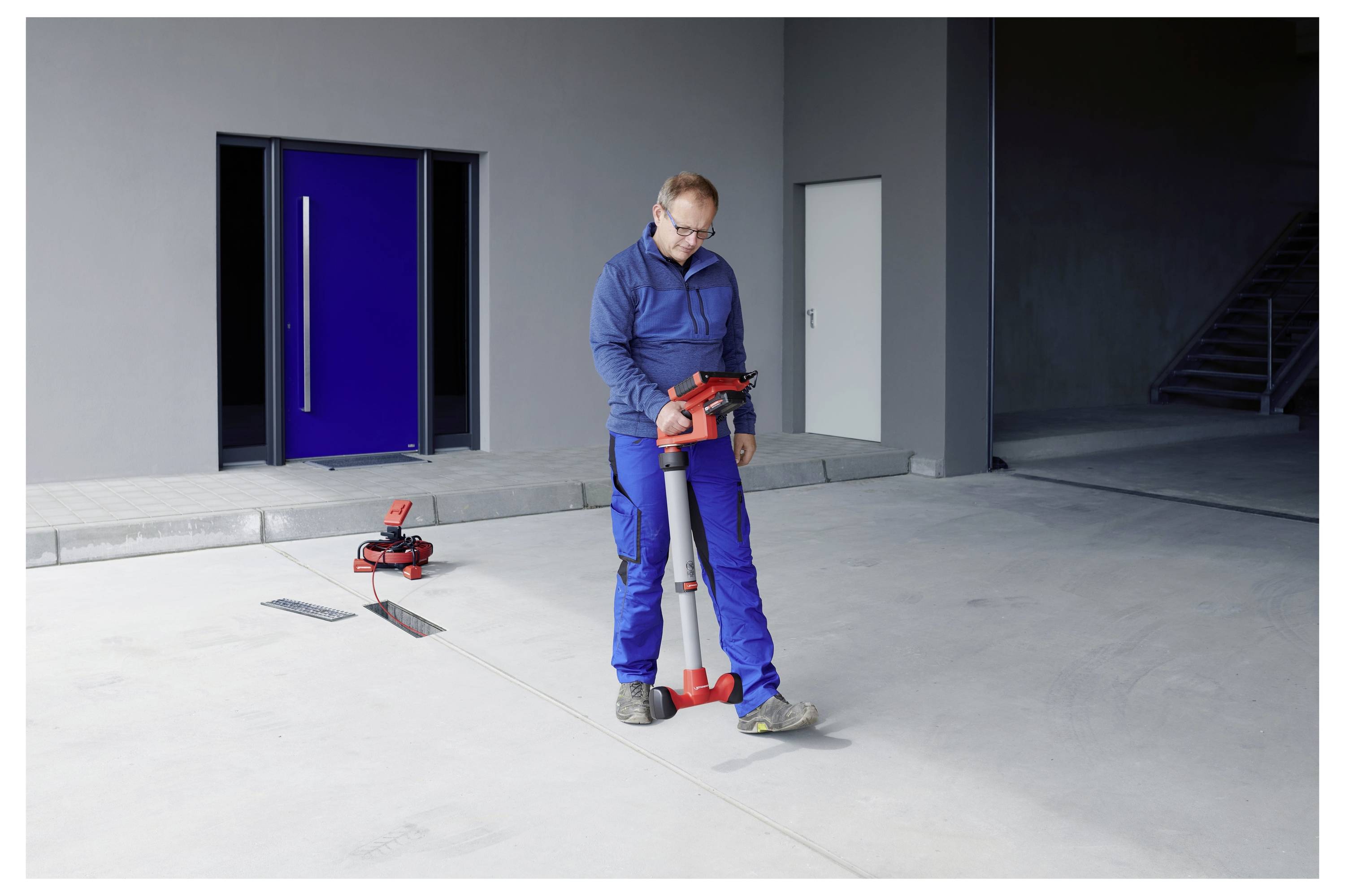 Un homme en bleu effectue une mesure de sol à l'aide d'un outil de nivellement laser rouge et gris dans un espace industriel avec une porte bleue en arrière-plan.