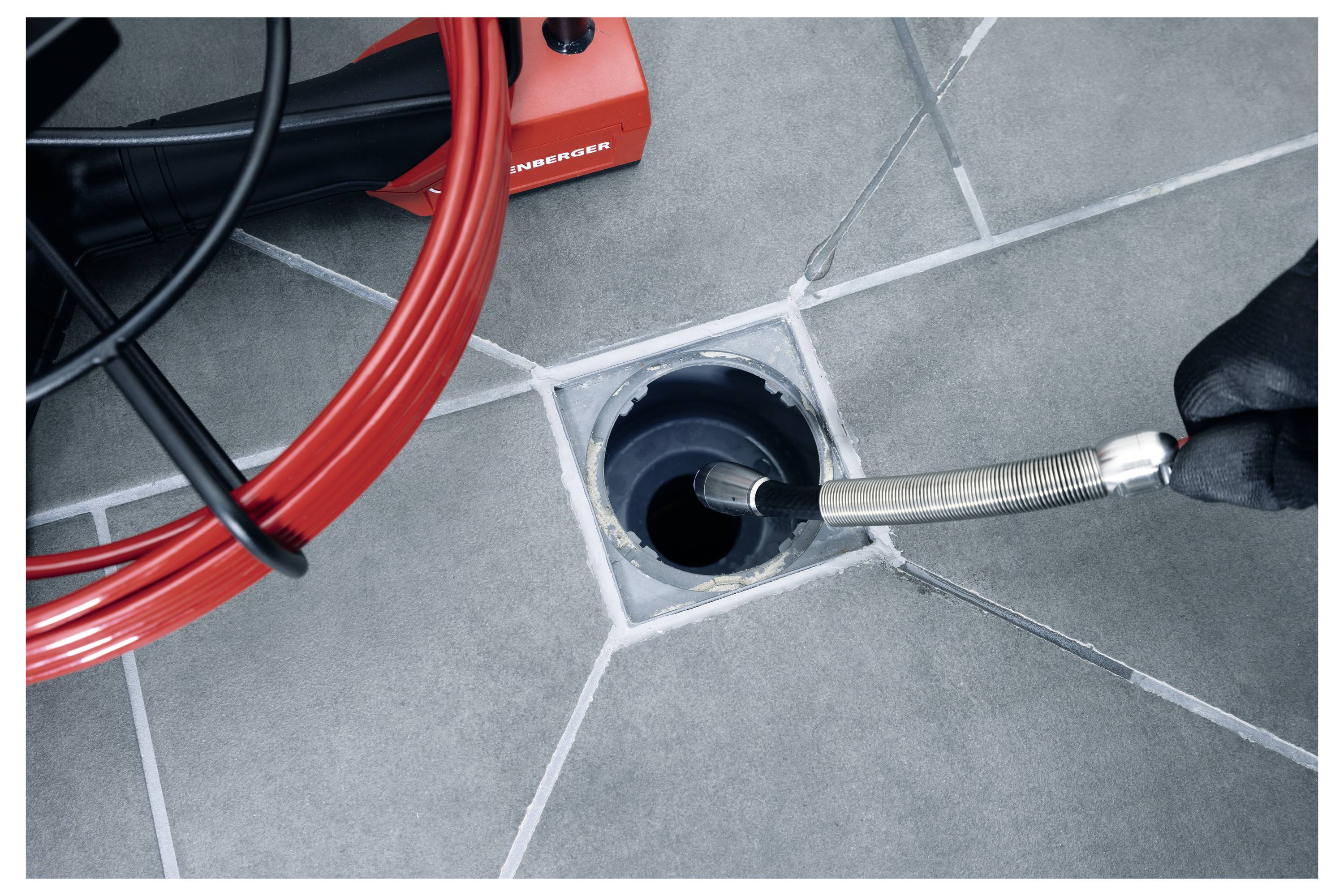 A drain cleaning machine with a red flexible rod is being inserted into a floor drain, illustrating plumbing maintenance.