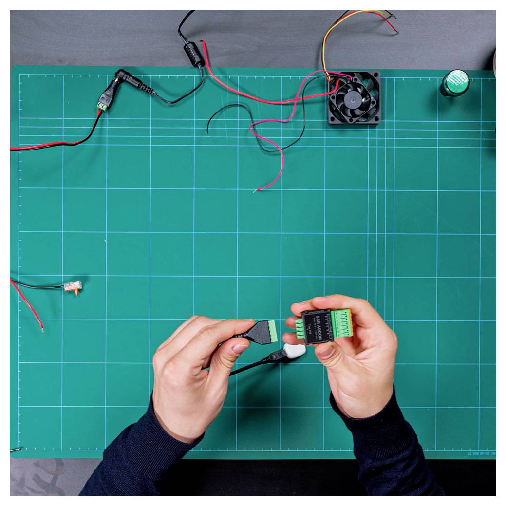 A person's hands on a green cutting mat connect electronic components with wires, with a fan and tools visible in the background.