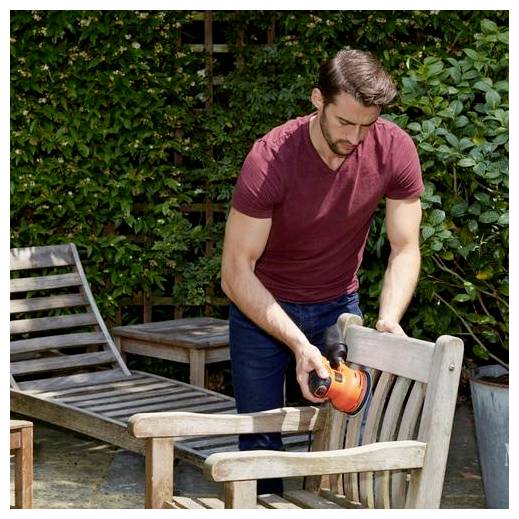 Une personne utilisant une ponceuse électrique sur une chaise en bois à l'extérieur. Des buissons verts en arrière-plan, le cadre indique des travaux de jardin ou un projet de bricolage.