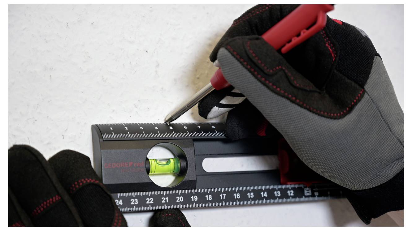 Gloved hands using a ruler with an integrated level to mark a measurement on a white surface, indicating a precise task setup.