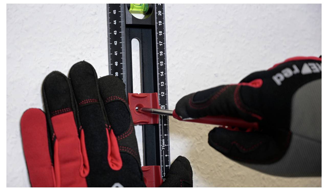 A person wearing gloves is using a screwdriver to adjust a tool with measurement markings on a vertical surface.