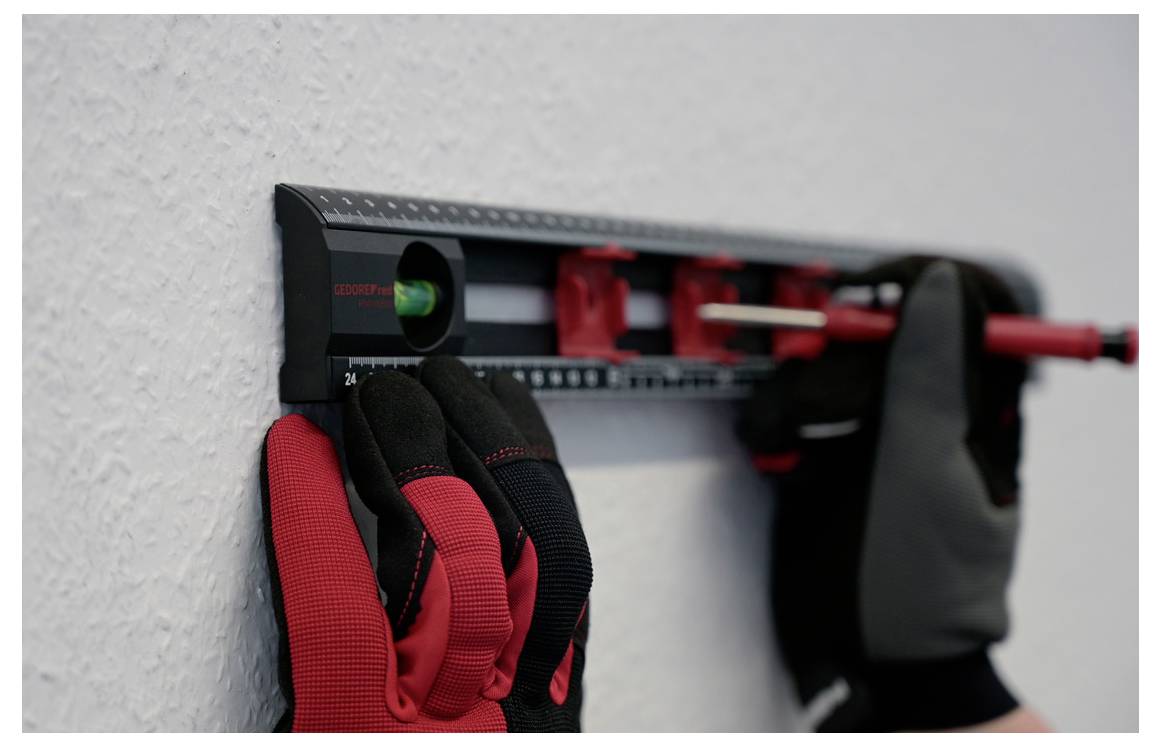 Hands wearing red and black gloves are using a red level tool to ensure a wall is straight.