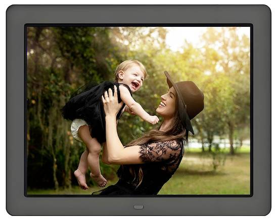 Une femme en robe noire et chapeau soulève un bébé souriant dans un parc, entourée d'arbres et d'une lumière chaude et douce.