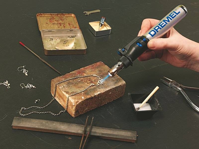Eine Person verwendet einen Dremel-Brenner zum Löten eines Metallanhängers an einer Halskette, umgeben von Schmuckherstellungswerkzeugen auf einem Tisch.