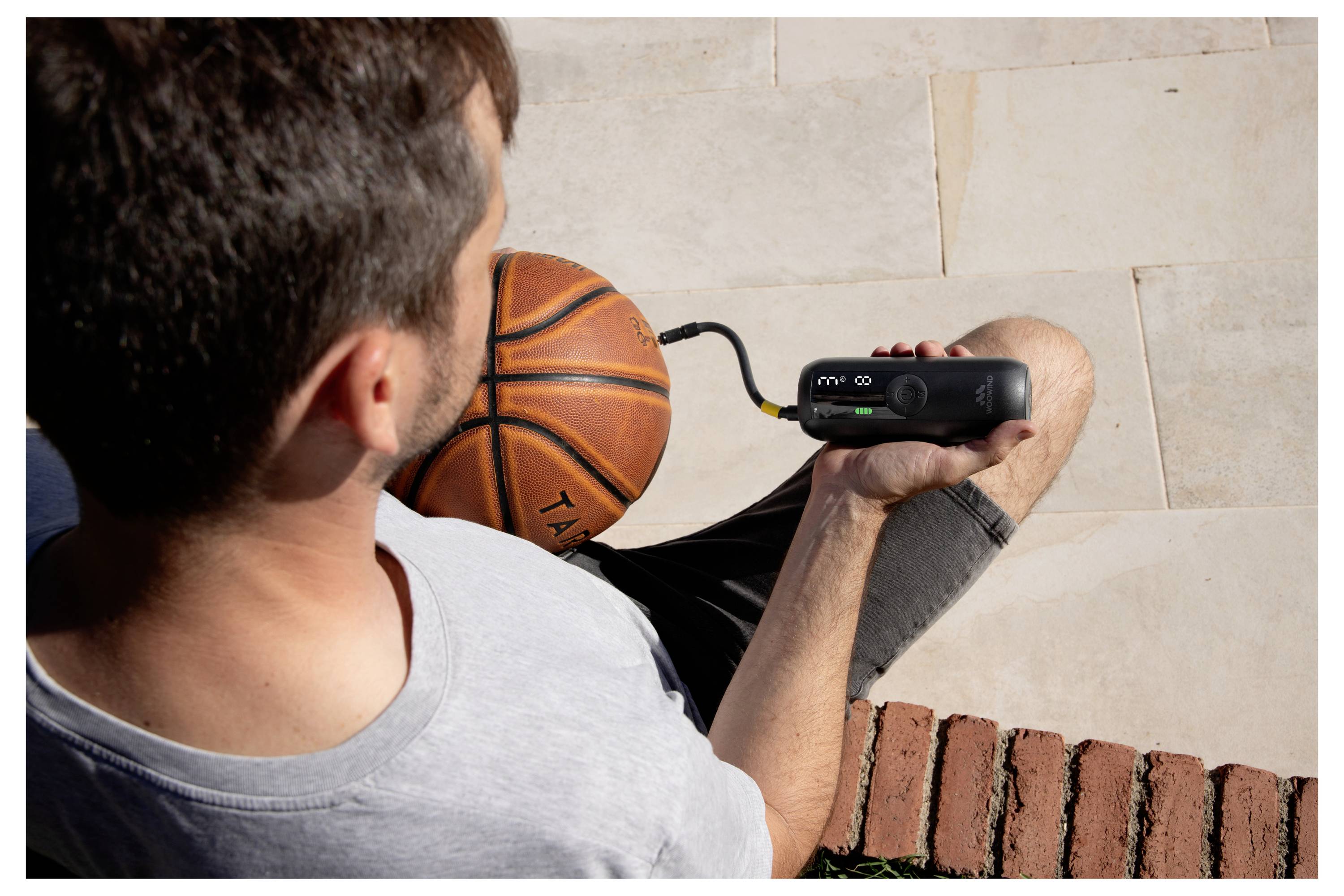 Draufsicht auf einen Mann, der eine tragbare Pumpe hält, während er einen Basketball auf gepflastertem Pflaster im Freien aufpumpt.