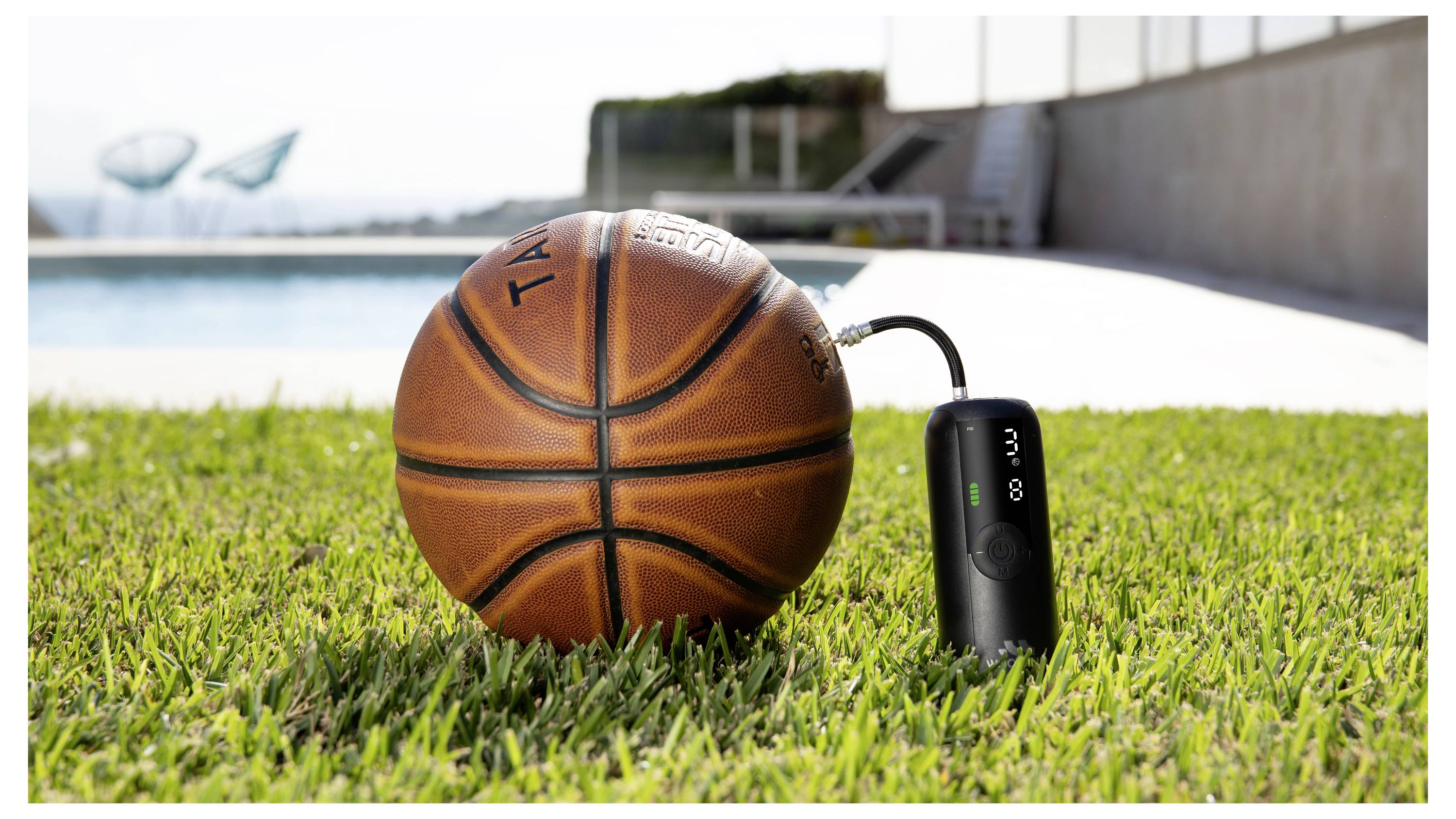 Ein Basketball liegt auf Gras neben einem kleinen schwarzen Gerät, mit einem Schwimmbad und einem Außenweg im Hintergrund.