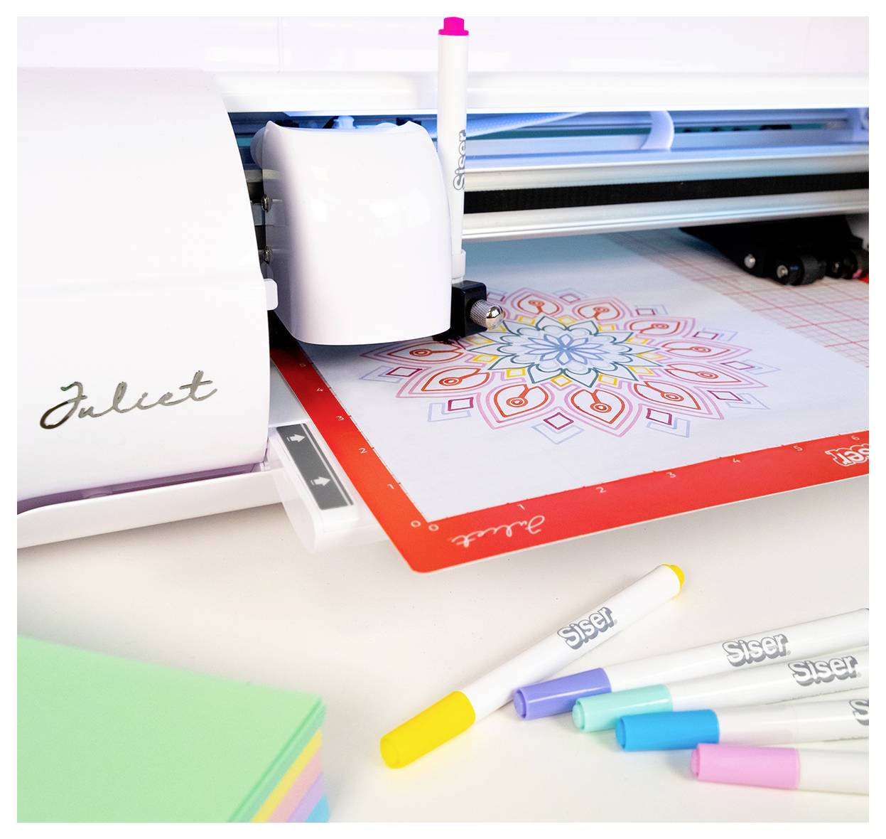 Une machine de découpe dessine un magnifique mandala coloré sur une feuille, entourée de marqueurs. La machine porte l'étiquette « Juliet ».