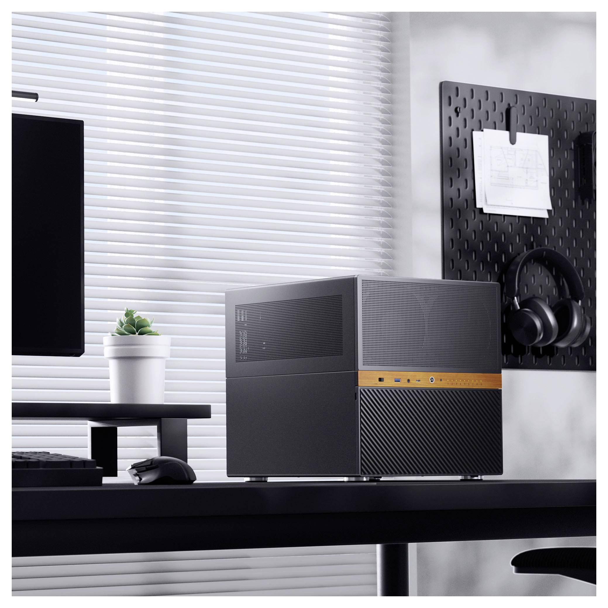 Un ordinateur de bureau moderne sur une table, avec une plante et un casque sur un panneau à chevilles mural, dans une pièce bien éclairée avec des stores.