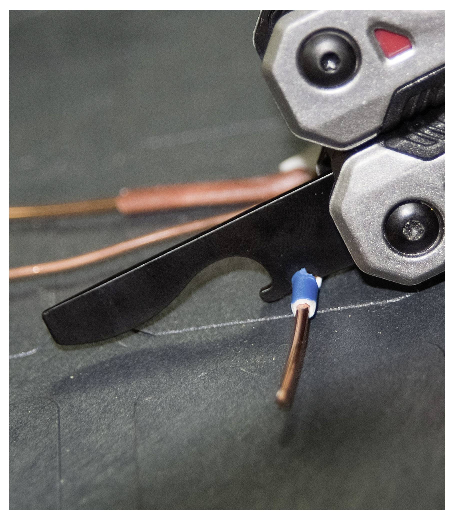 Close-up of a wire stripper tool gripping an insulated copper wire, ready for stripping. The tool is metallic with visible screws.
