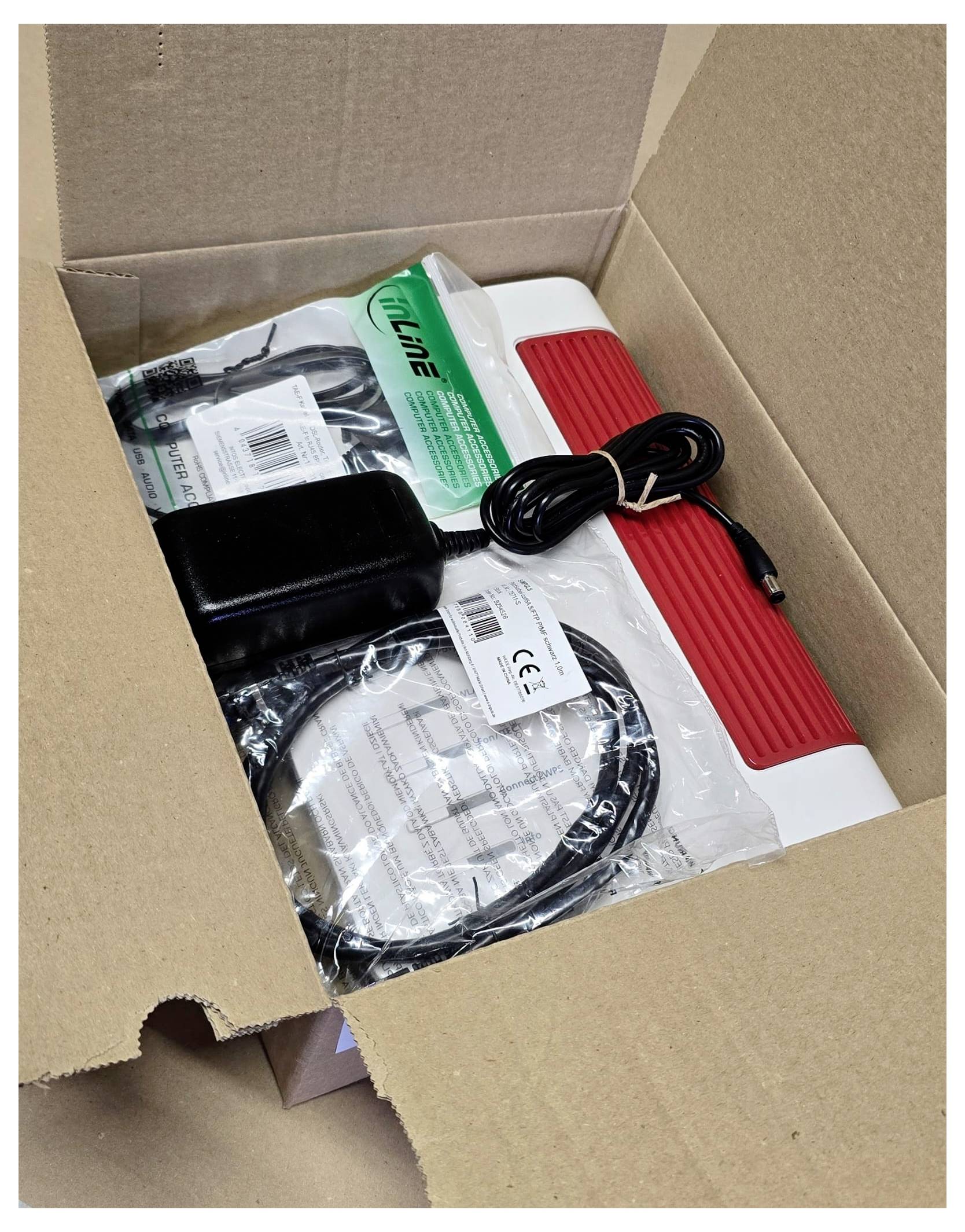 Eine geöffnete Kartonbox enthält ein elektronisches Gerät mit einem roten Griff, aufgerollte Kabel, einen Stromadapter und eine Bedienungsanleitung.