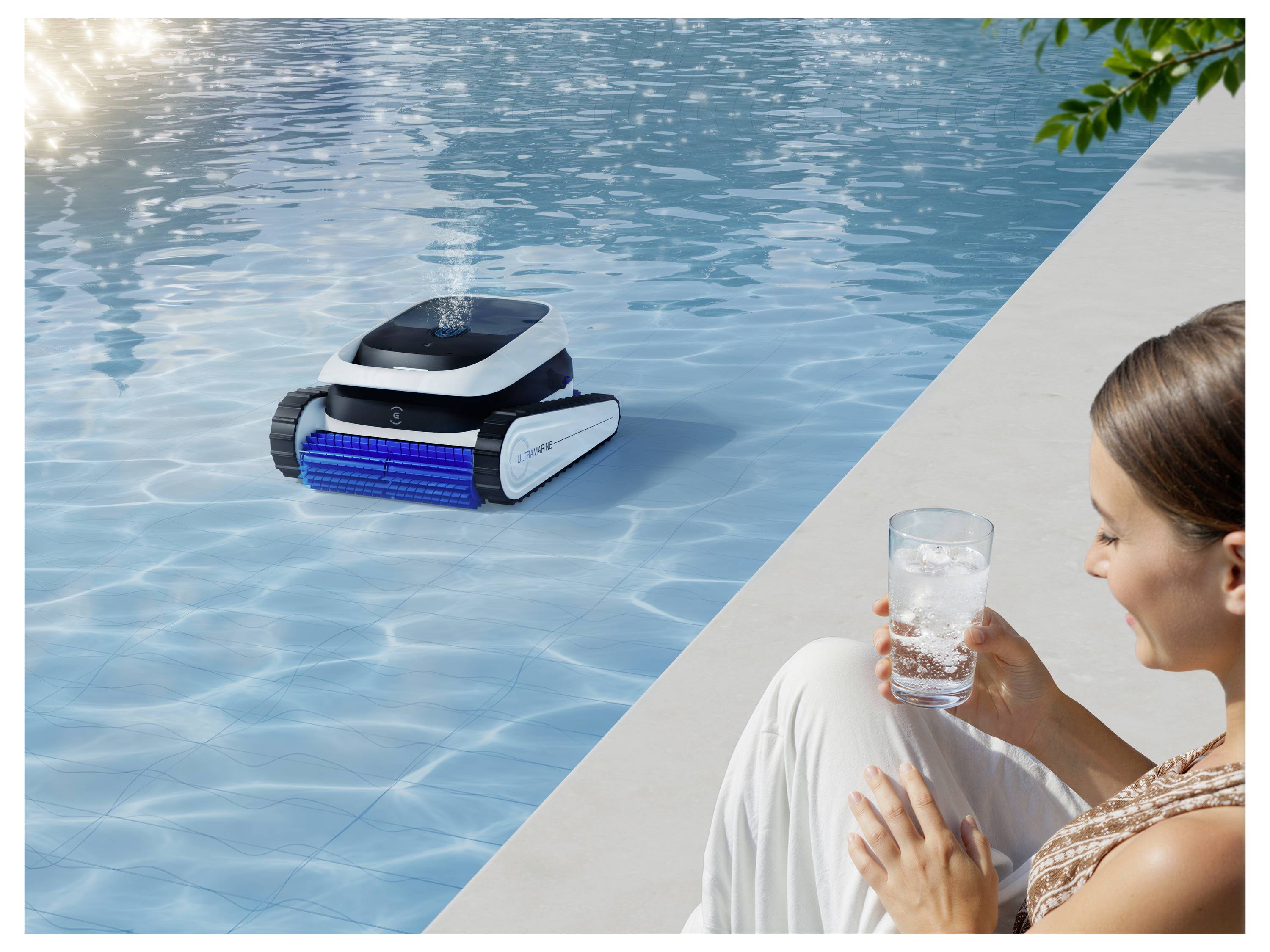 Une femme est assise près d'une piscine en tenant un verre, regardant un robot nettoyeur de piscine se déplacer dans l'eau.