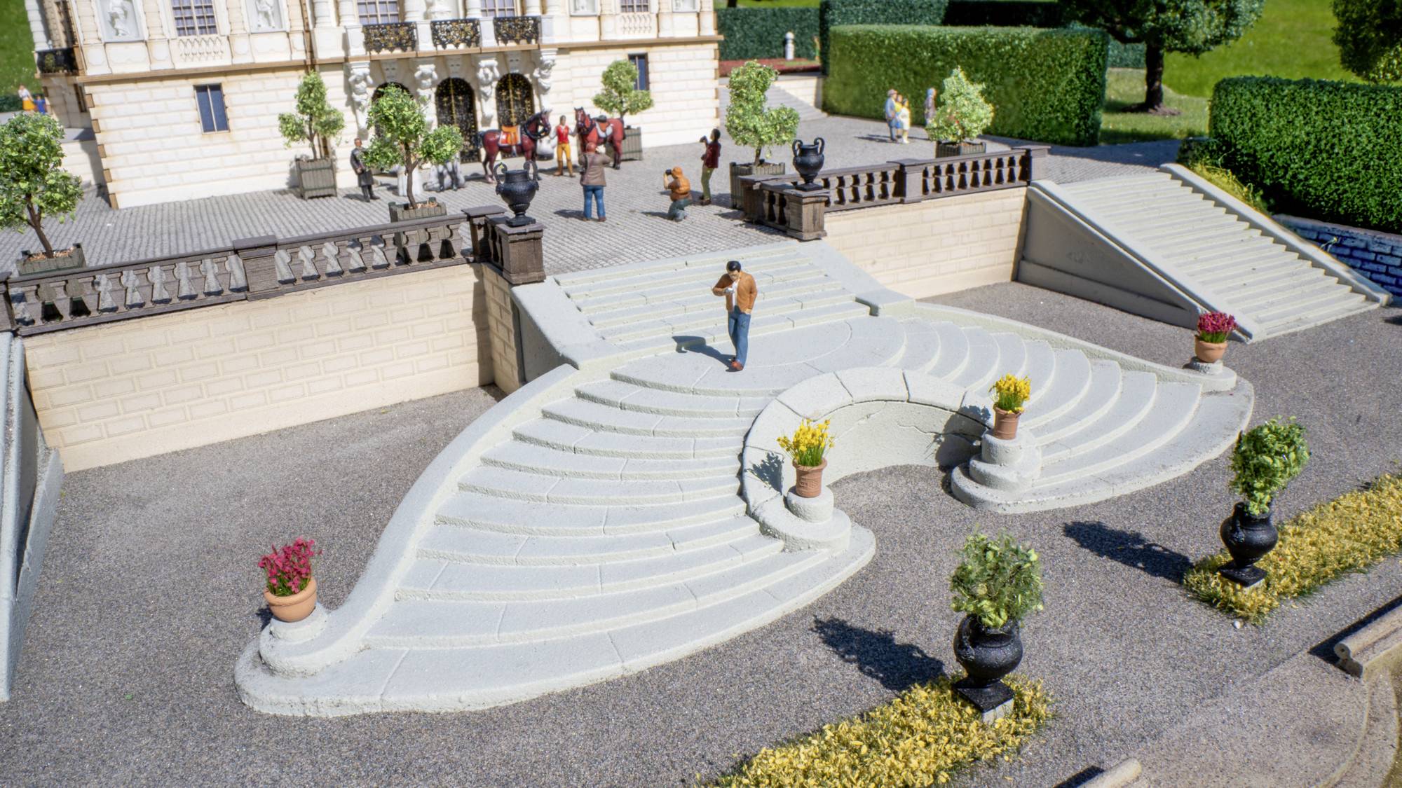 Modèle miniature de terrasse extérieure avec des marches blanches courbes et élancées, des fleurs en pot et de minuscules figurines devant un grand bâtiment.