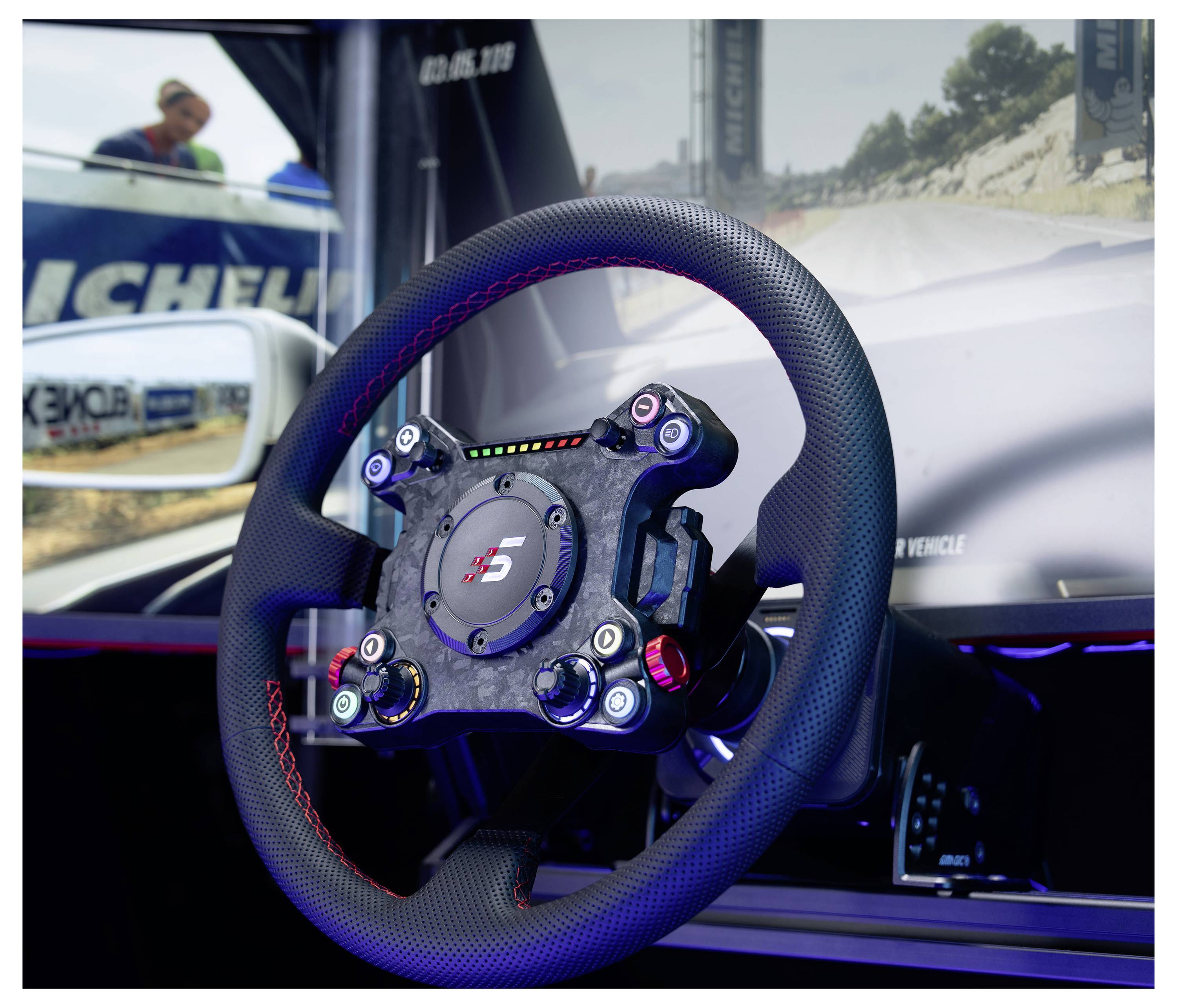 Gros plan d'un volant de voiture de course monté dans un simulateur ou un cockpit d'exposition, avec une scène de course floue et la marque Michelin en arrière-plan.