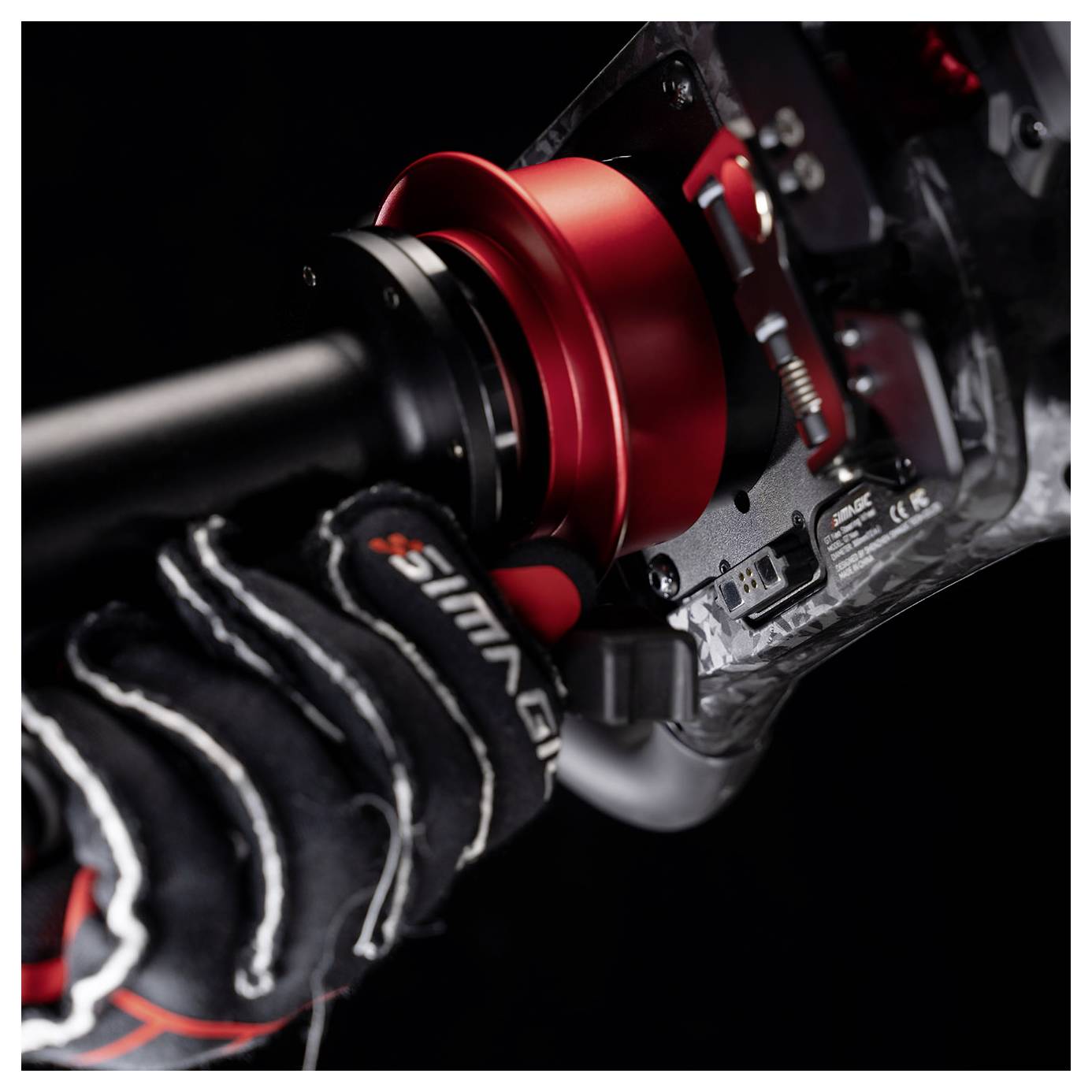 Gros plan d'une main gantée utilisant un volant de course rouge fixé à un cockpit en fibre de carbone ou à un panneau de commande de simulateur.