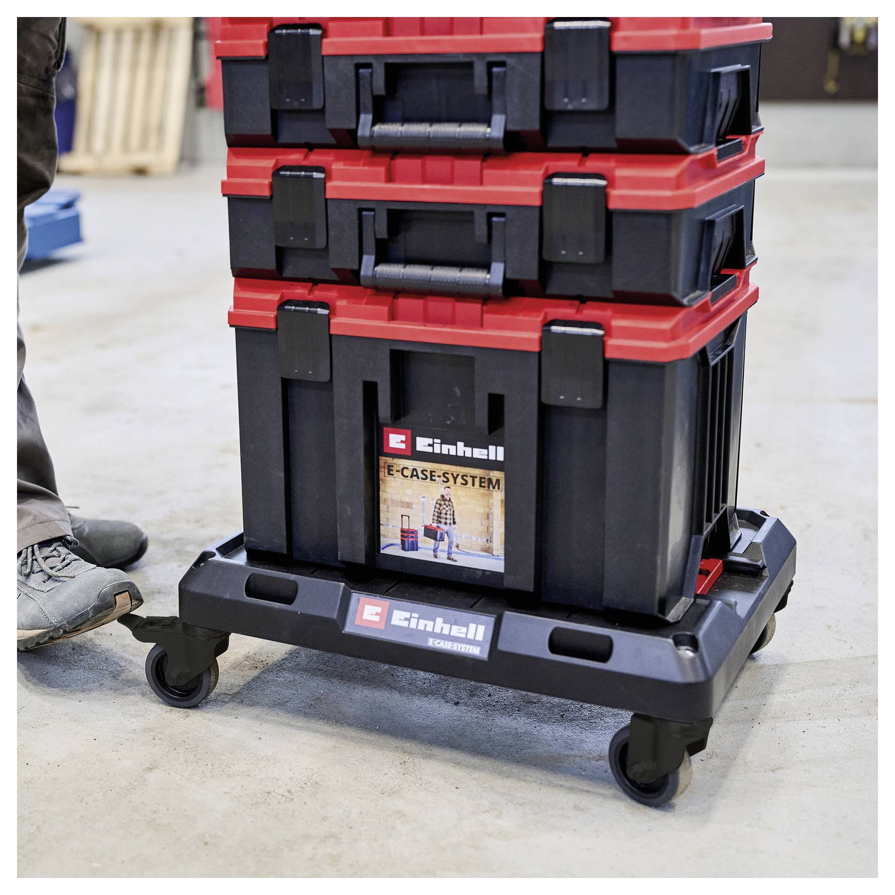 Des boîtes de rangement d'outils Einhell empilées sur un chariot à roulettes dans un atelier, avec la chaussure d'une personne partiellement visible à gauche.