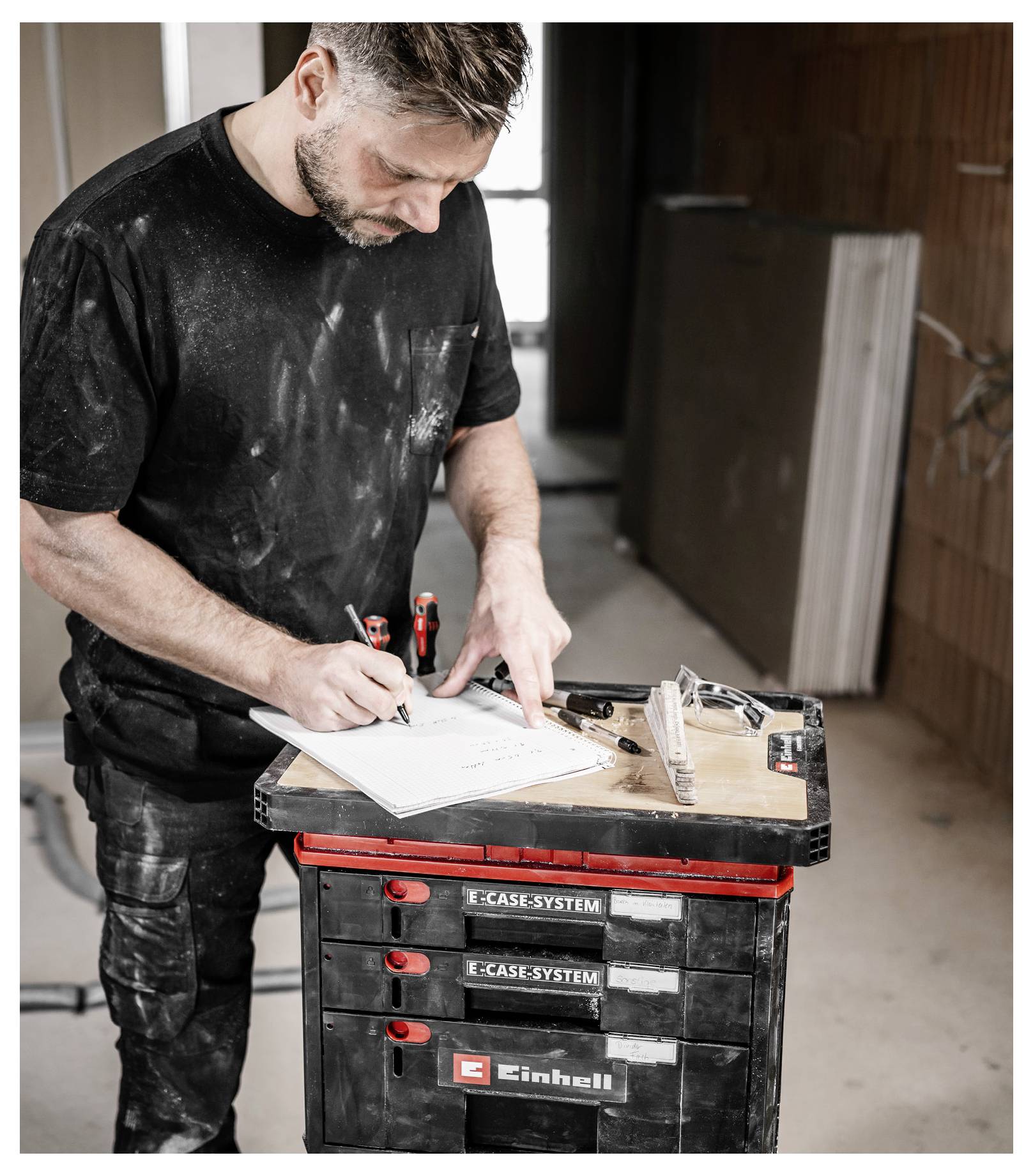 Homme prenant des mesures et des notes sur un presse-papiers à côté d'une mallette à outils dans une zone de travail de rénovation.