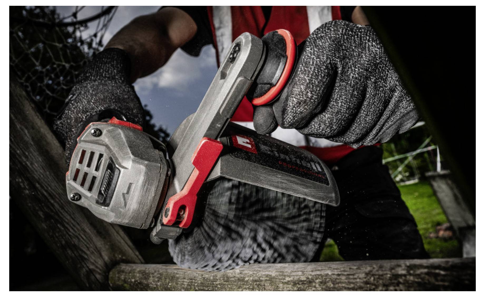Gros plan d'une personne portant des gants utilisant un outil électrique rouge et noir sur une surface en bois à l'extérieur.