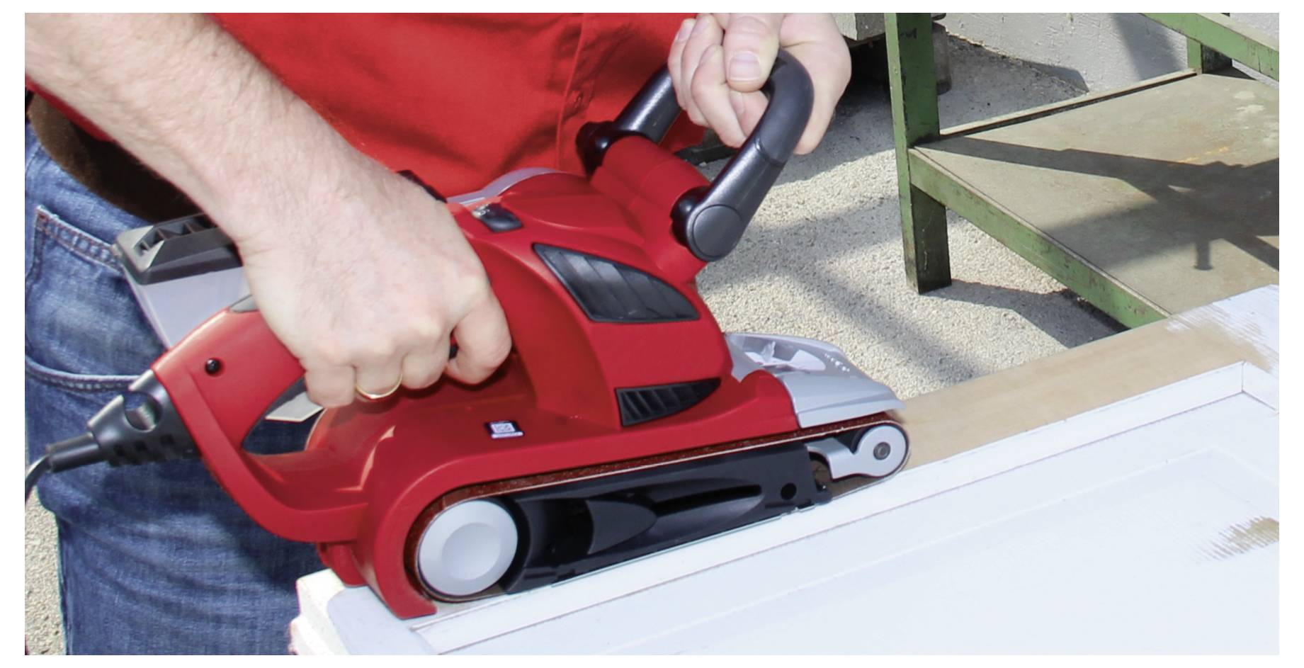 Close-up of a person using a red electric hand planer to smooth the edge of a wooden board outdoors.