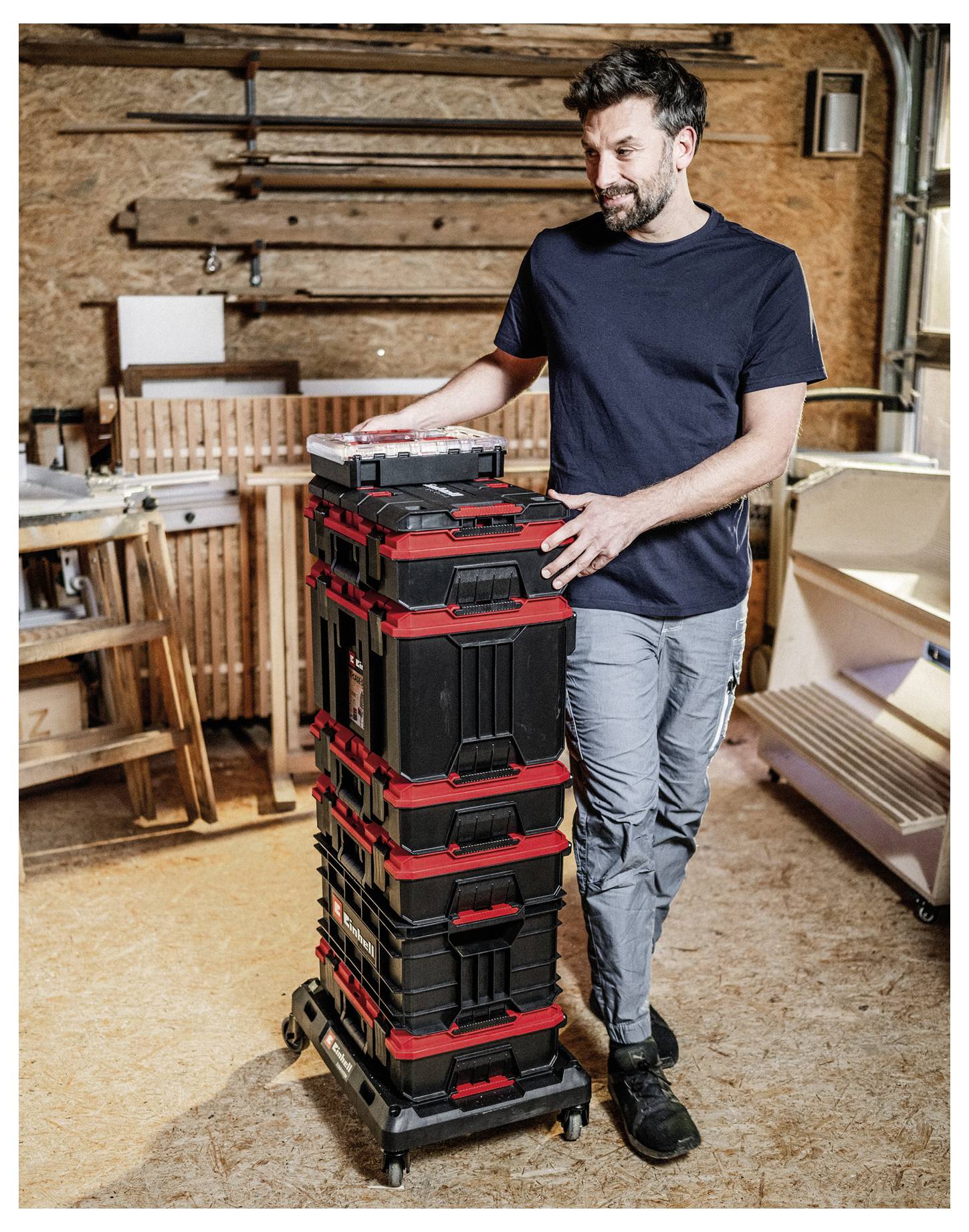 Man in a workshop standing beside a wheeled stack of black-and-red tool storage boxes, with wood panels and furniture in the background.