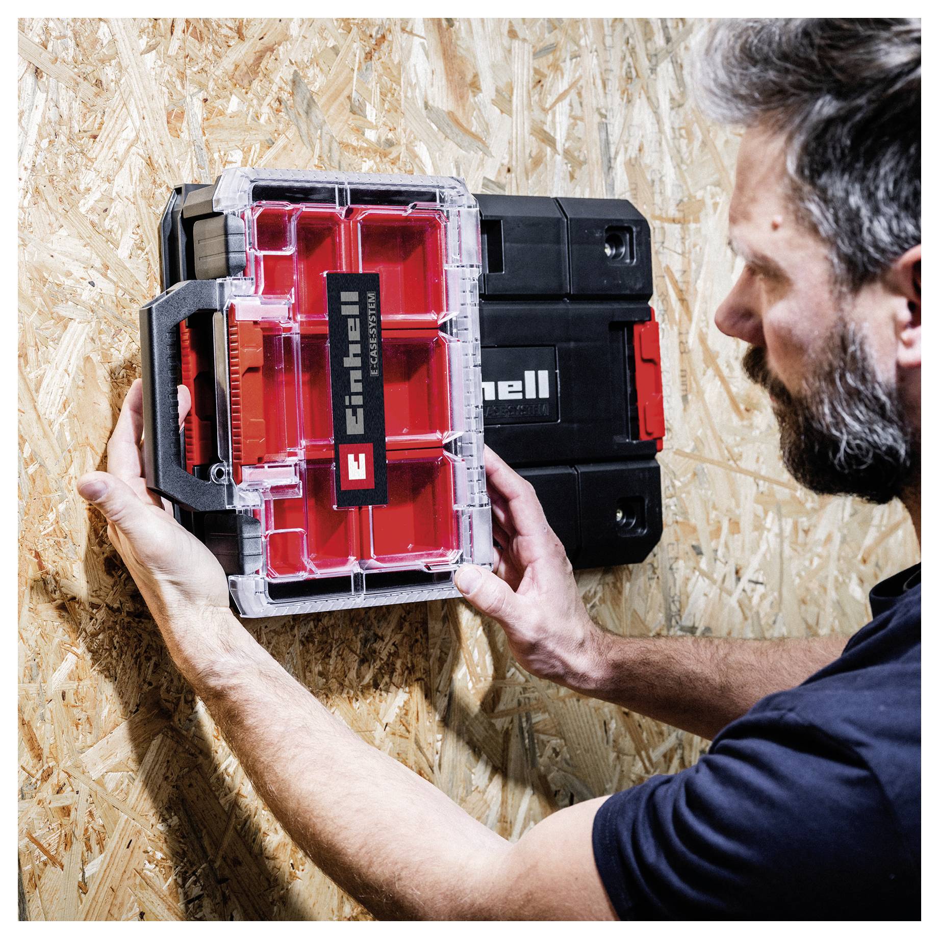 Man mounting a red and black Einhell battery organizer on an OSB plywood wall using a hanging bracket.