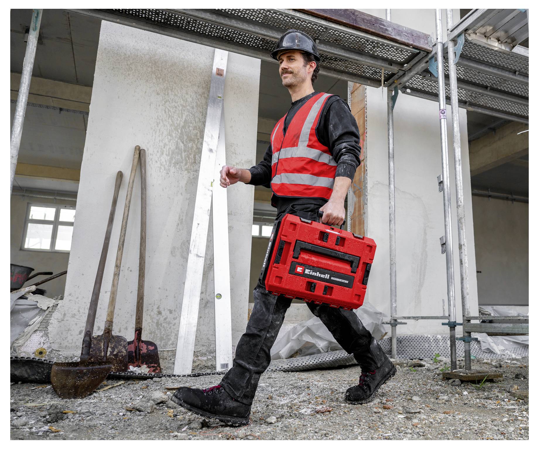 Ouvrier du bâtiment portant un casque de protection et un gilet de sécurité marchant avec une mallette à outils rouge sur un chantier de construction.