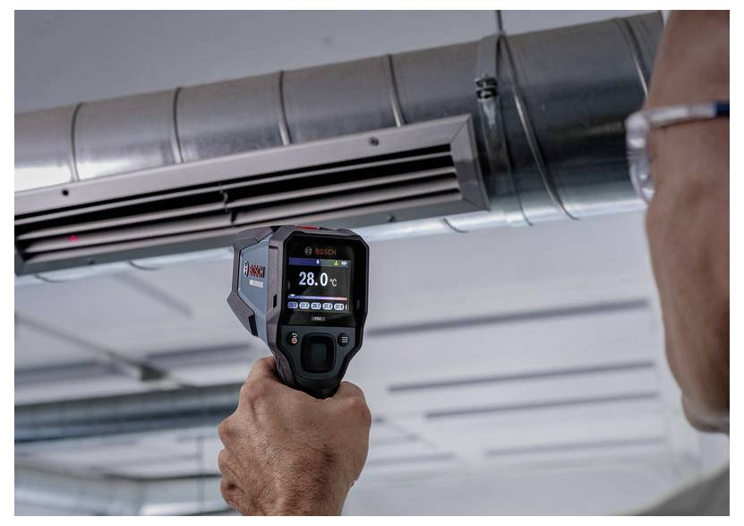 Une personne utilise une caméra thermique pour mesurer la température sur la tuyauterie de climatisation au plafond, l'appareil affichant 28,0°C.