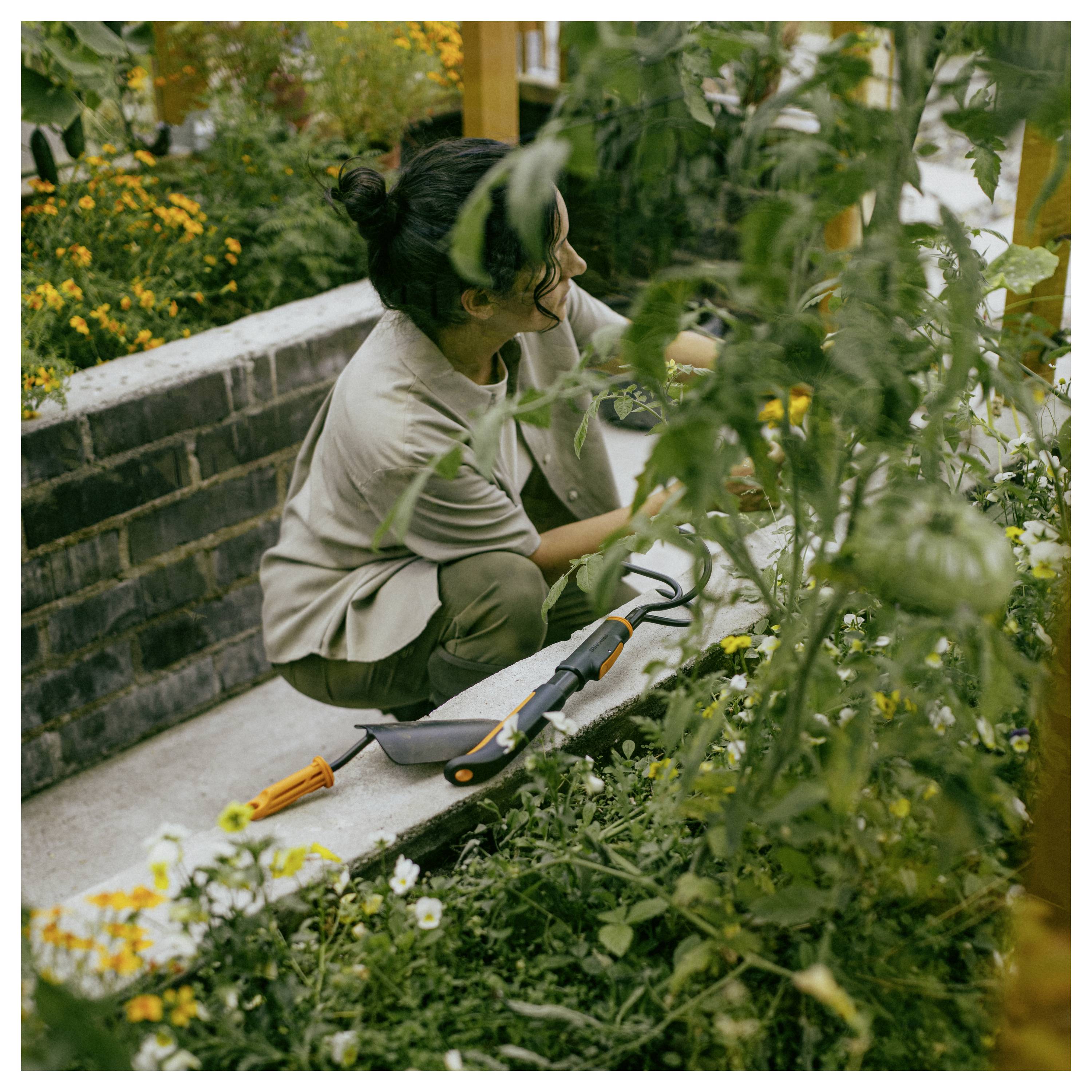 Une femme s'accroupit dans un parterre de jardin, s'occupant des plantes avec des outils à main à côté d'un mur de jardinière en brique.