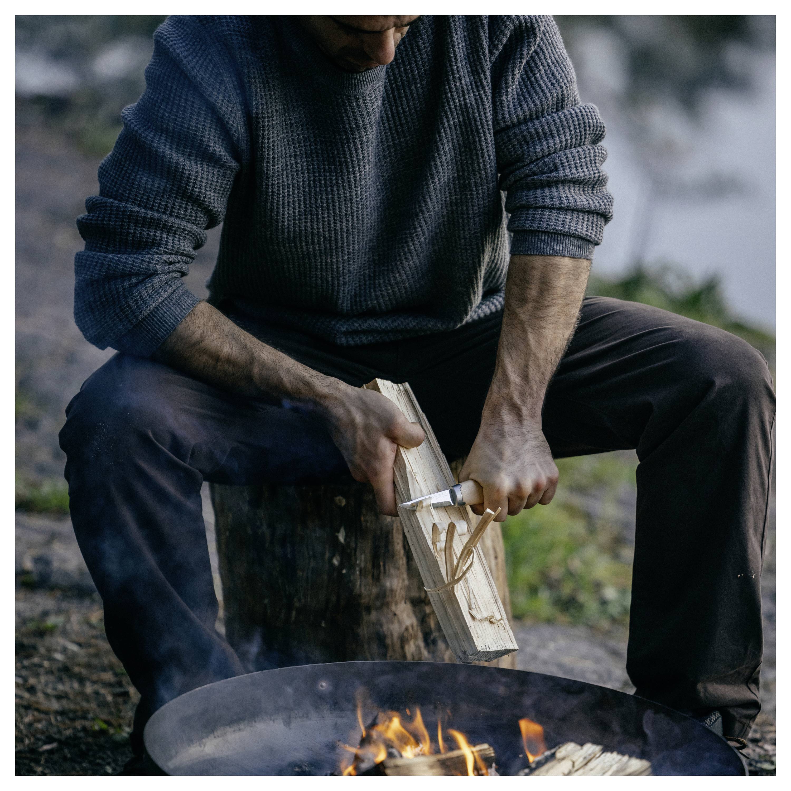 Une personne assise près d'un feu de camp, utilisant un couteau pour enlever des éclats de bois d'une bûche et les jeter dans le foyer.