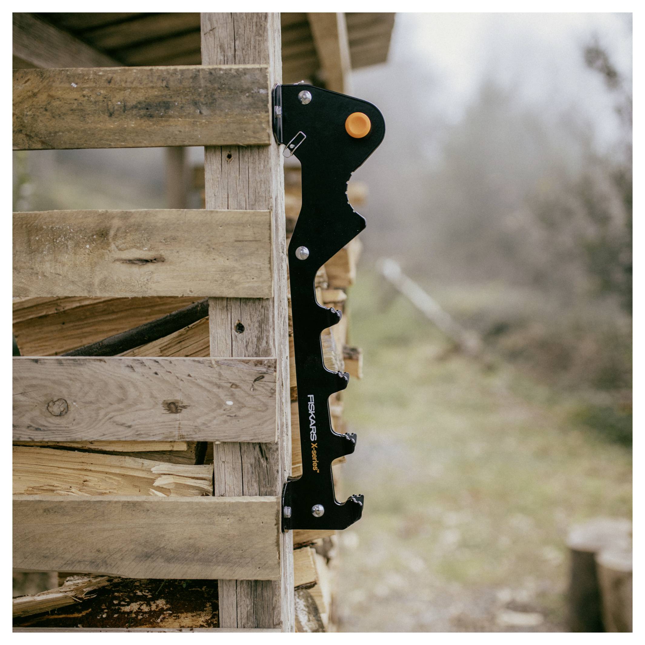 Séparateur de bois FISKARS noir attaché à une caisse de bois empilé à l'extérieur, avec un bouton orange et une cour floue en arrière-plan.
