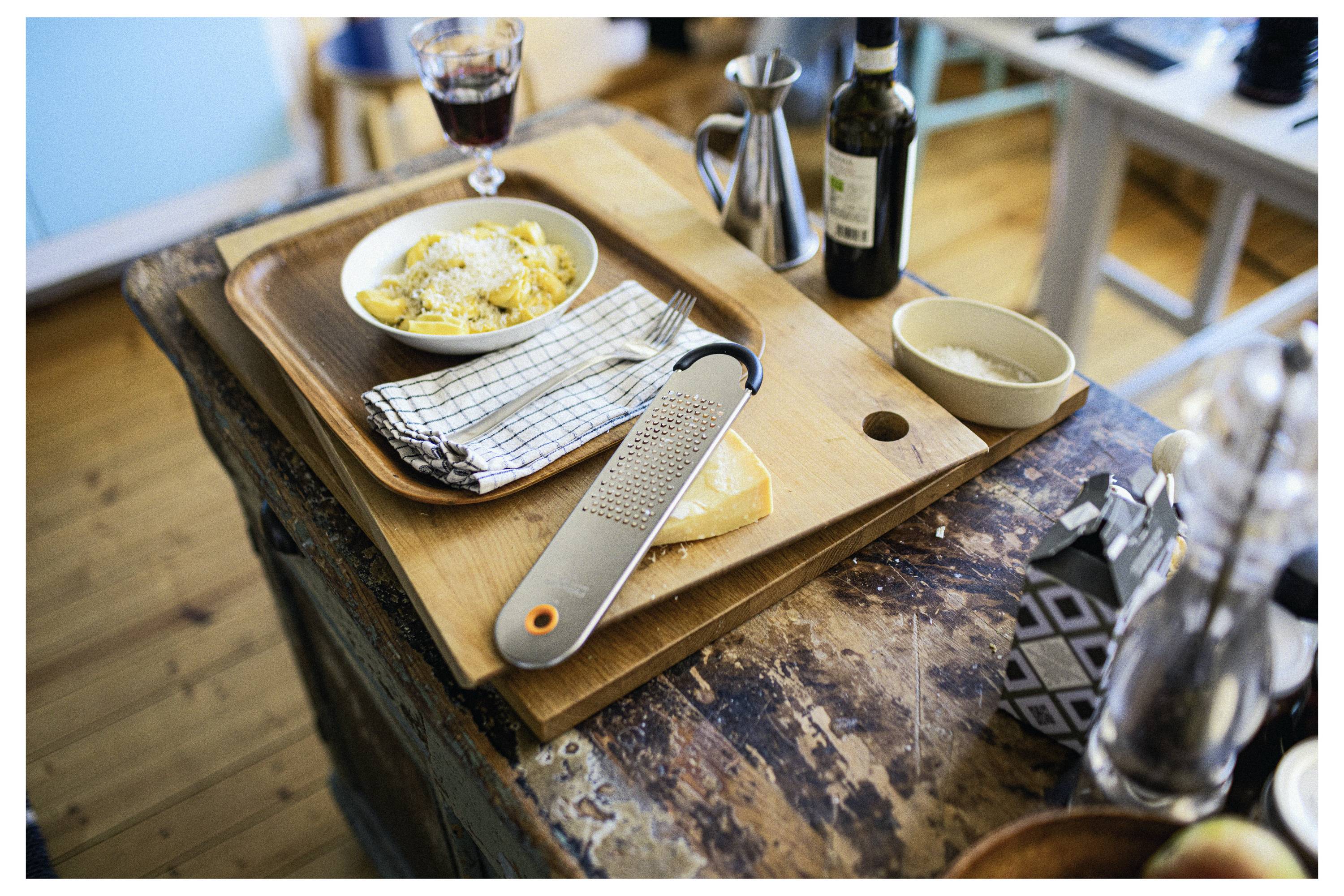 Tischgedeck mit einer Reibe, Käse, einer Schüssel mit geriebenem Käse, gefalteter Serviette, einem Glas Rotwein, einer Olivenölflasche und einem kleinen Krug auf Holzschneidebrettern.