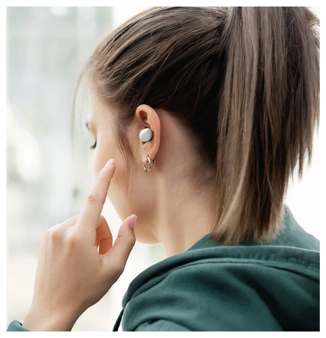 Personne portant un écouteur sans fil et une boucle d'oreille, touchant l'oreille en se tournant avec un sweat-shirt bleu-vert.