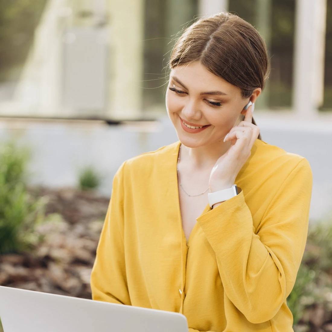 Femme souriante vêtue d'un chemisier jaune utilisant des écouteurs tout en regardant un ordinateur portable à l'extérieur.