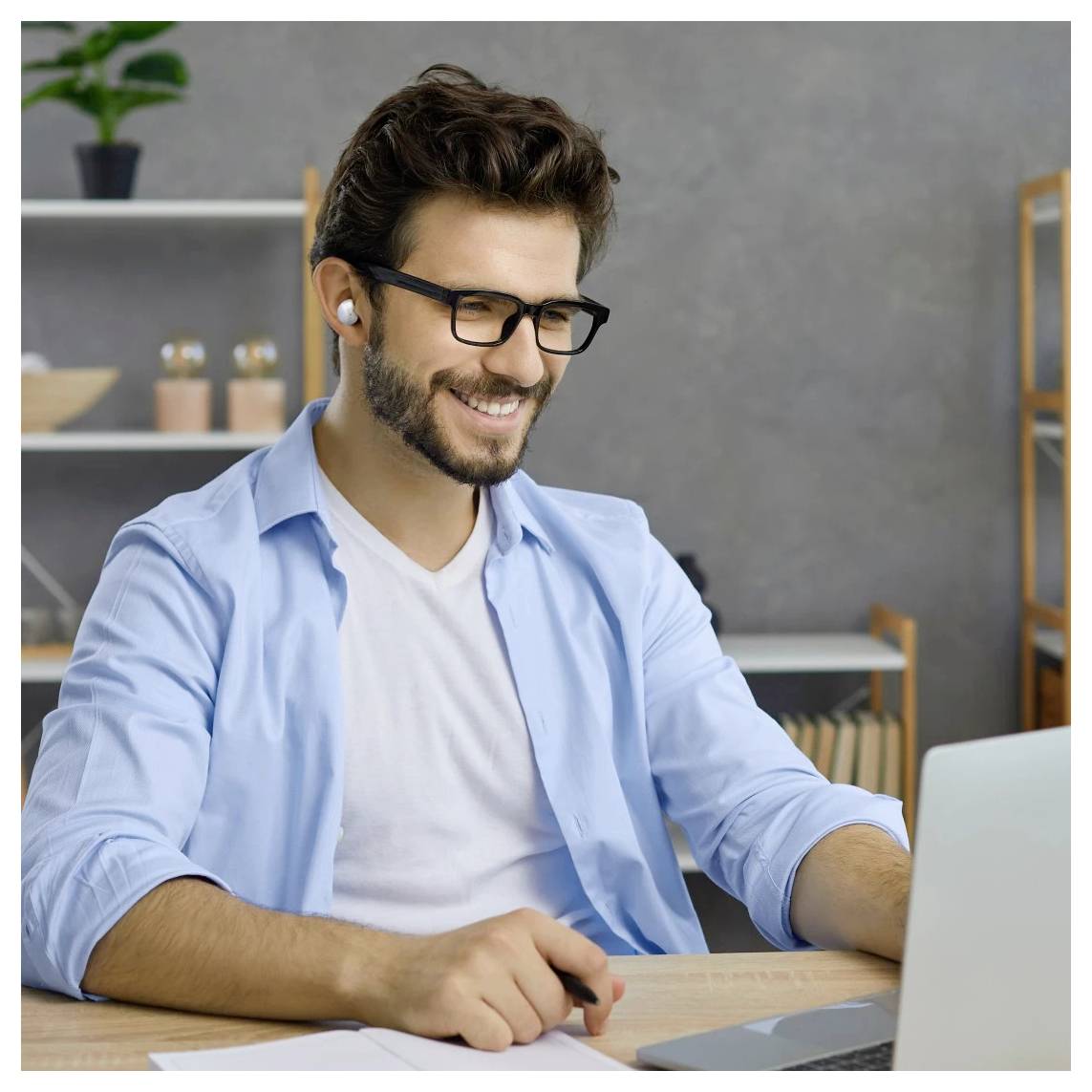 Smiling man with glasses and earbuds works on a laptop at a desk in a home office.