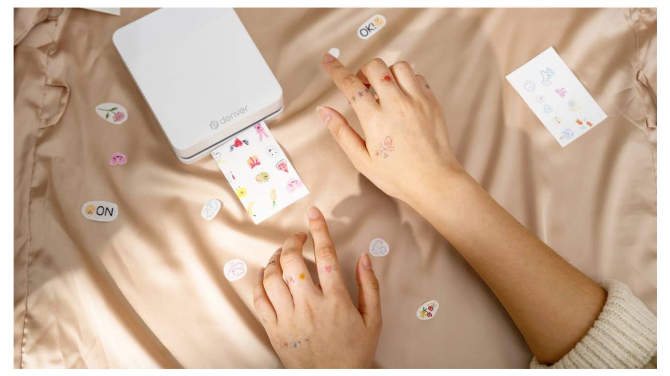 Deux mains utilisent une petite imprimante photo Canon blanche pour imprimer une feuille d'autocollants colorée sur un lit, avec des autocollants décoratifs éparpillés autour.