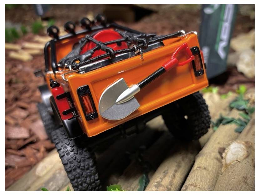 Camion RC tout-terrain escaladant des bûches, portant une pelle à manche rouge sur son panneau de chargement arrière.