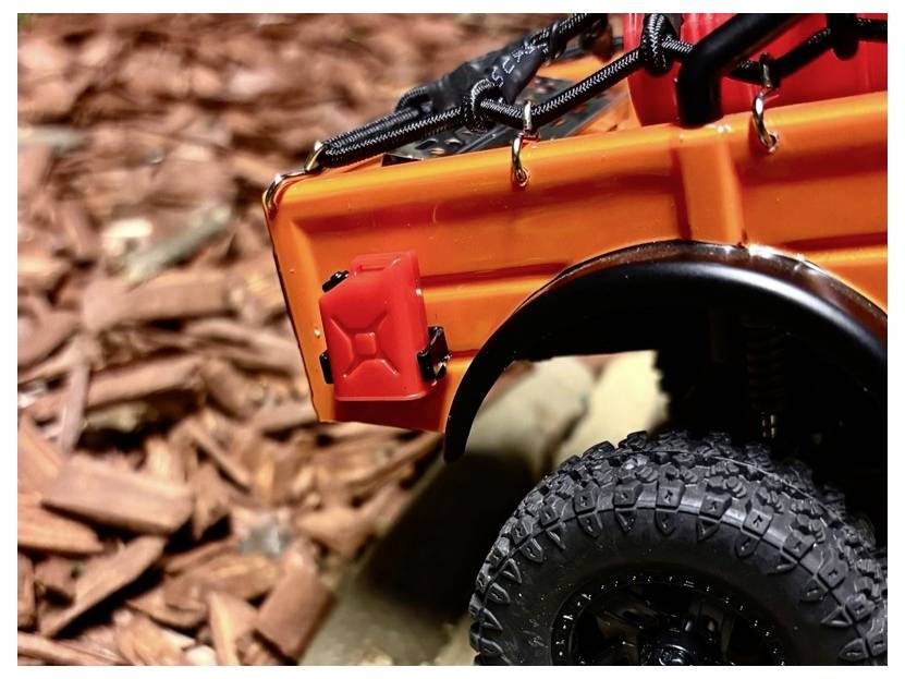 Gros plan d'un véhicule tout-terrain jouet orange avec un pneu noir et un bidon d'essence rouge, présenté sur des copeaux de bois et une pierre.