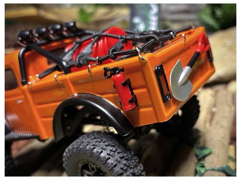 Camion RC tout-terrain orange avec pneus surdimensionnés et équipement de toit, présenté sur un terrain accidenté dans une vue de profil en gros plan.