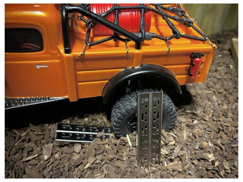 Orange Offroad-RC-Lastwagen mit Dachfrachtgitter und Ersatzreifen hinten, geparkt auf Holzspänen mit Metallstützteilen neben dem Rad.