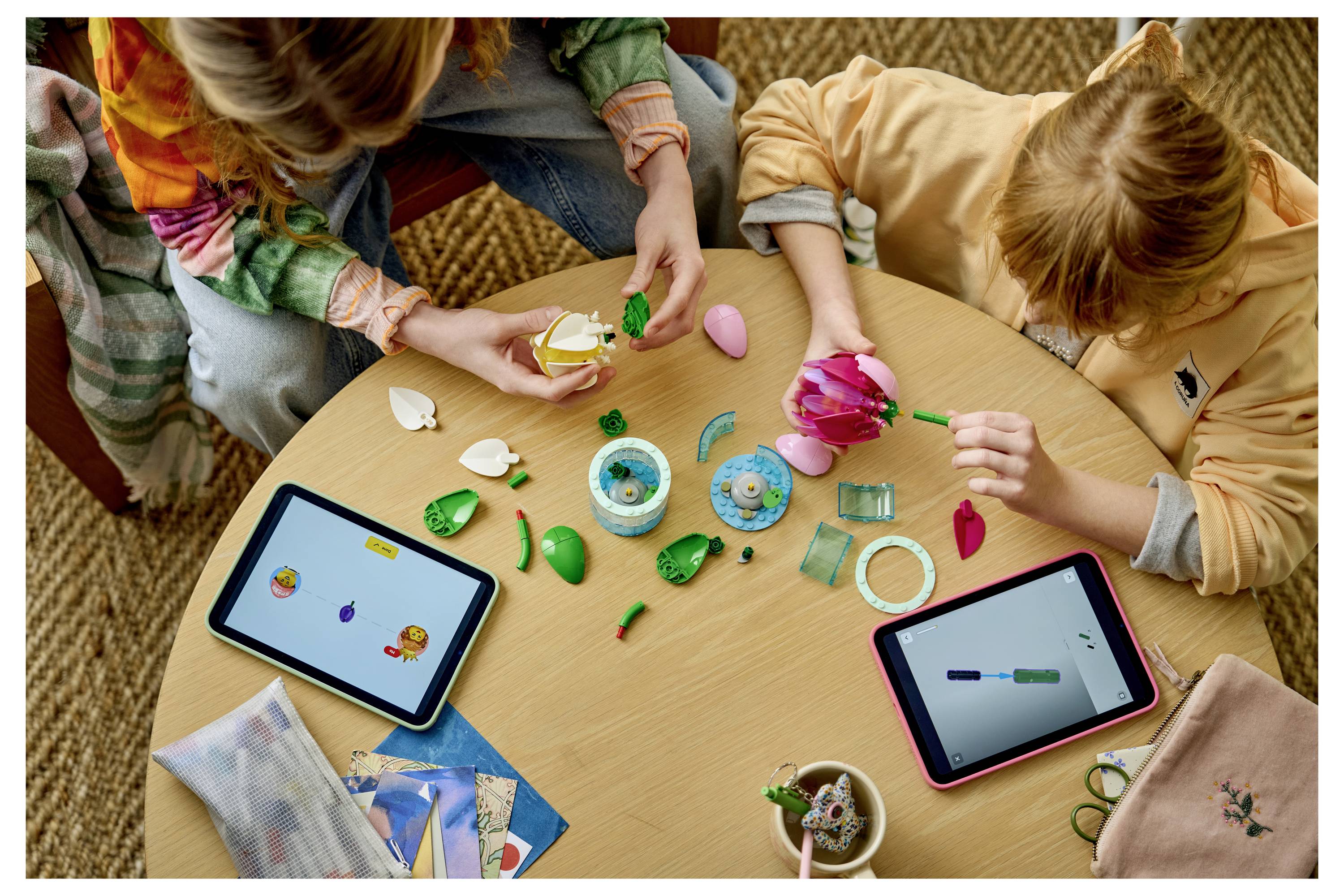 Les enfants sont assis autour d'une table en train d'assembler des pièces d'artisanat colorées et des perles tout en utilisant deux tablettes, ce qui suggère une activité créative collaborative ou un jeu.