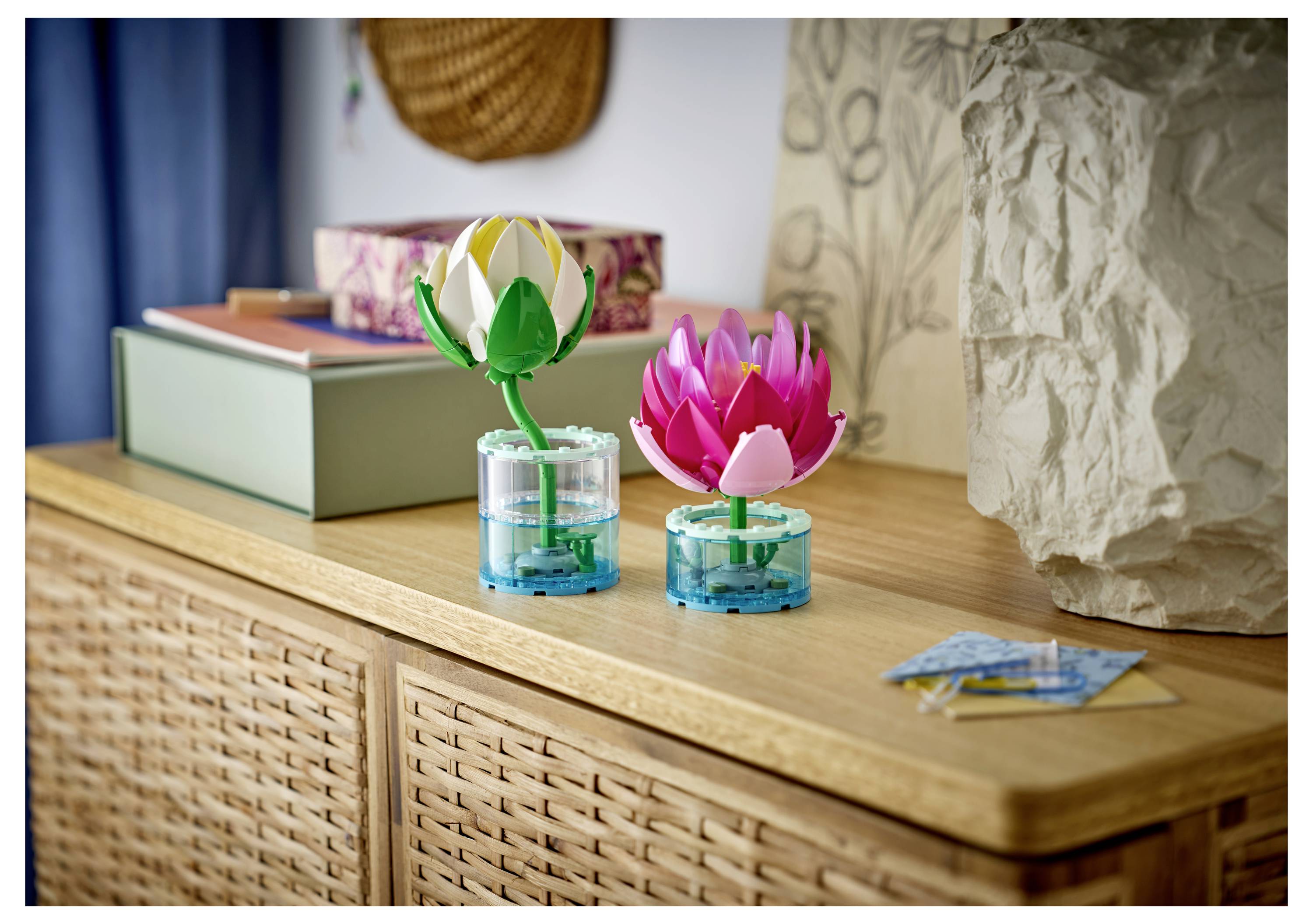 Deux petites fleurs de lotus de style LEGO en bleu sont posées dans des pots sur une commode en bois à côté d'une sculpture en papier et d'objets décoratifs.