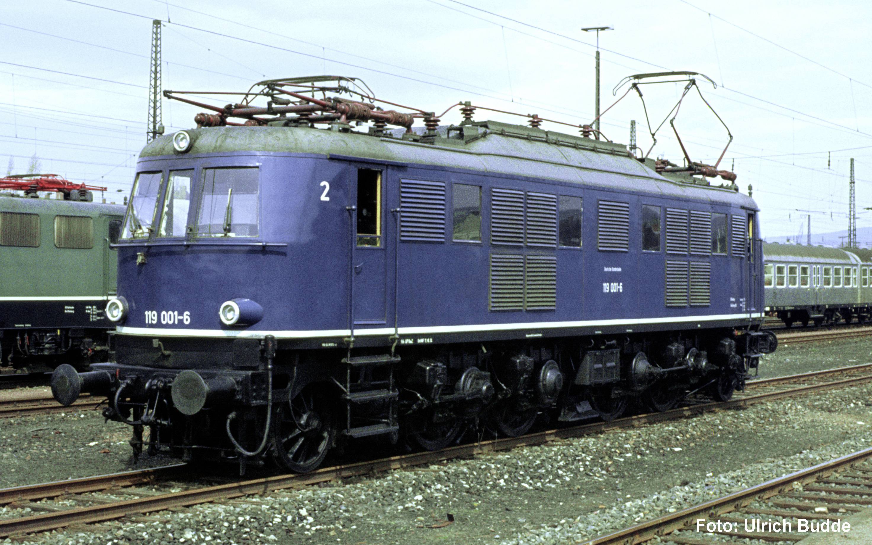 Locomotive électrique bleue numérotée 119 001-6 sur les rails de chemin de fer dans une cour de triage, avec des lignes électriques aériennes et d'autres voitures de train en arrière-plan.