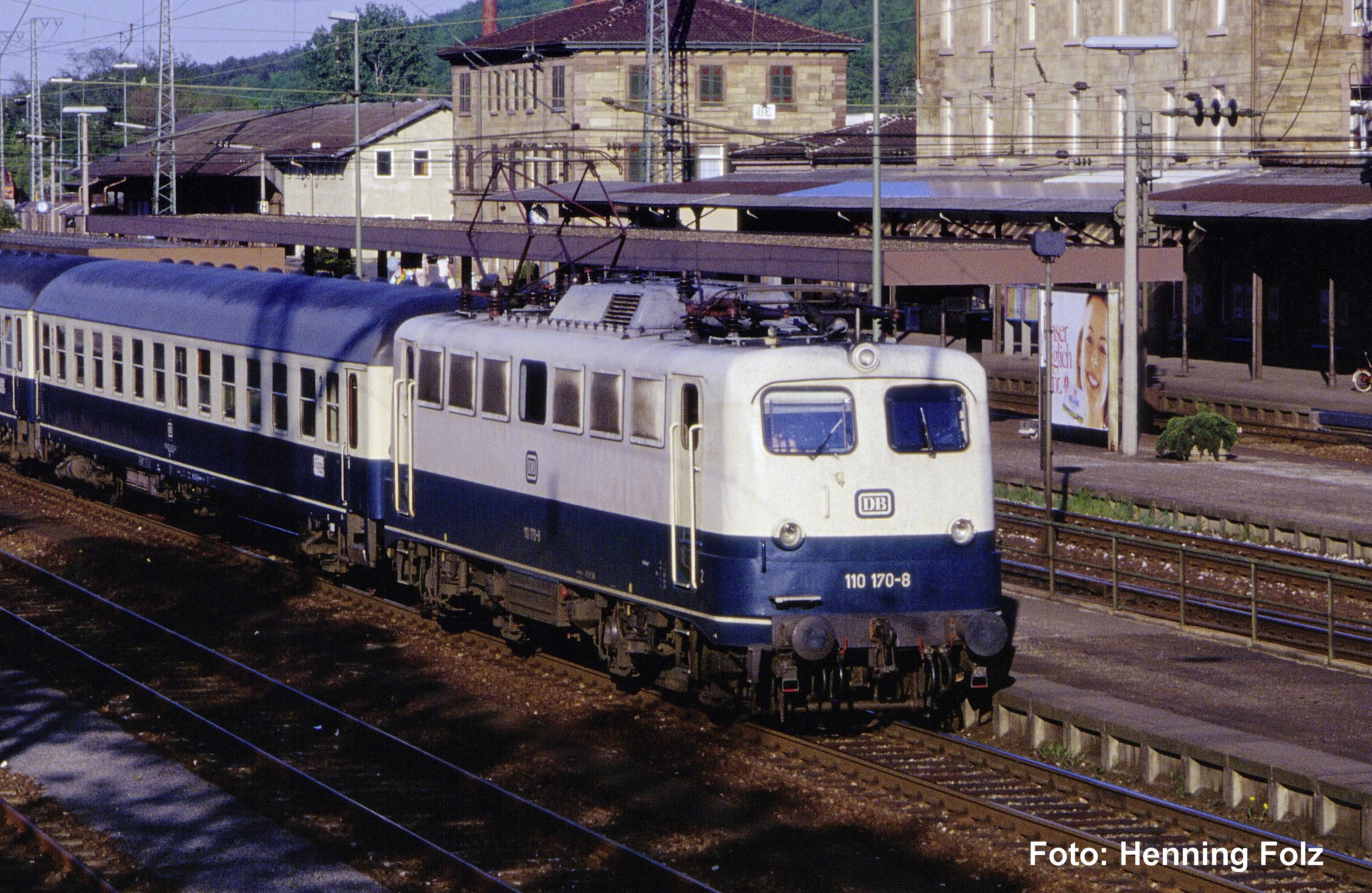 Train de passagers électrique bleu et crème à quai en gare, avec des fils aériens et des bâtiments de gare en arrière-plan.
